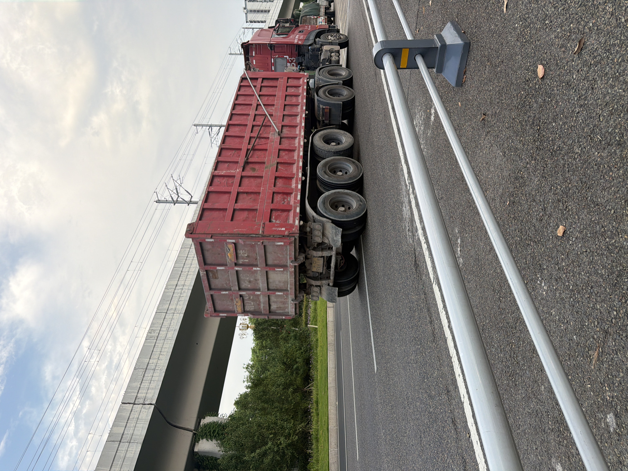 A red dump truck with a large cargo bed is driving on an asphalt road in Jiangjin, China. The truck is in the right lane of a multi-lane road, with a white line marking the lane. In the background, there is an elevated concrete structure, possibly a bridge or overpass, with electrical wires running overhead. Green trees and vegetation are visible on the left side of the road, and some buildings can be seen in the distance on the right. The sky is overcast with some clouds. A metal guardrail is in the foreground of the image.