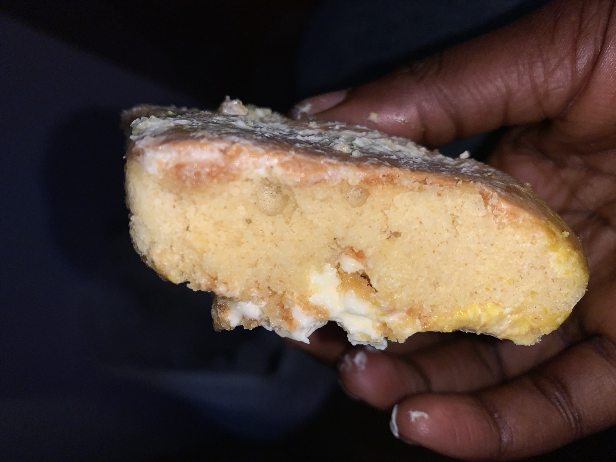 A slice of cake with white frosting and some reddish-orange streaks is held in a person's hand. The cake appears to be dense with a slightly crumbly texture. The frosting is thick and unevenly applied, with some of it having dripped onto the cake's edge and the hand holding it. The background is dark and indistinct, suggesting a low-light environment. No specific location in Gura Top, Nigeria can be determined from the visual information provided.