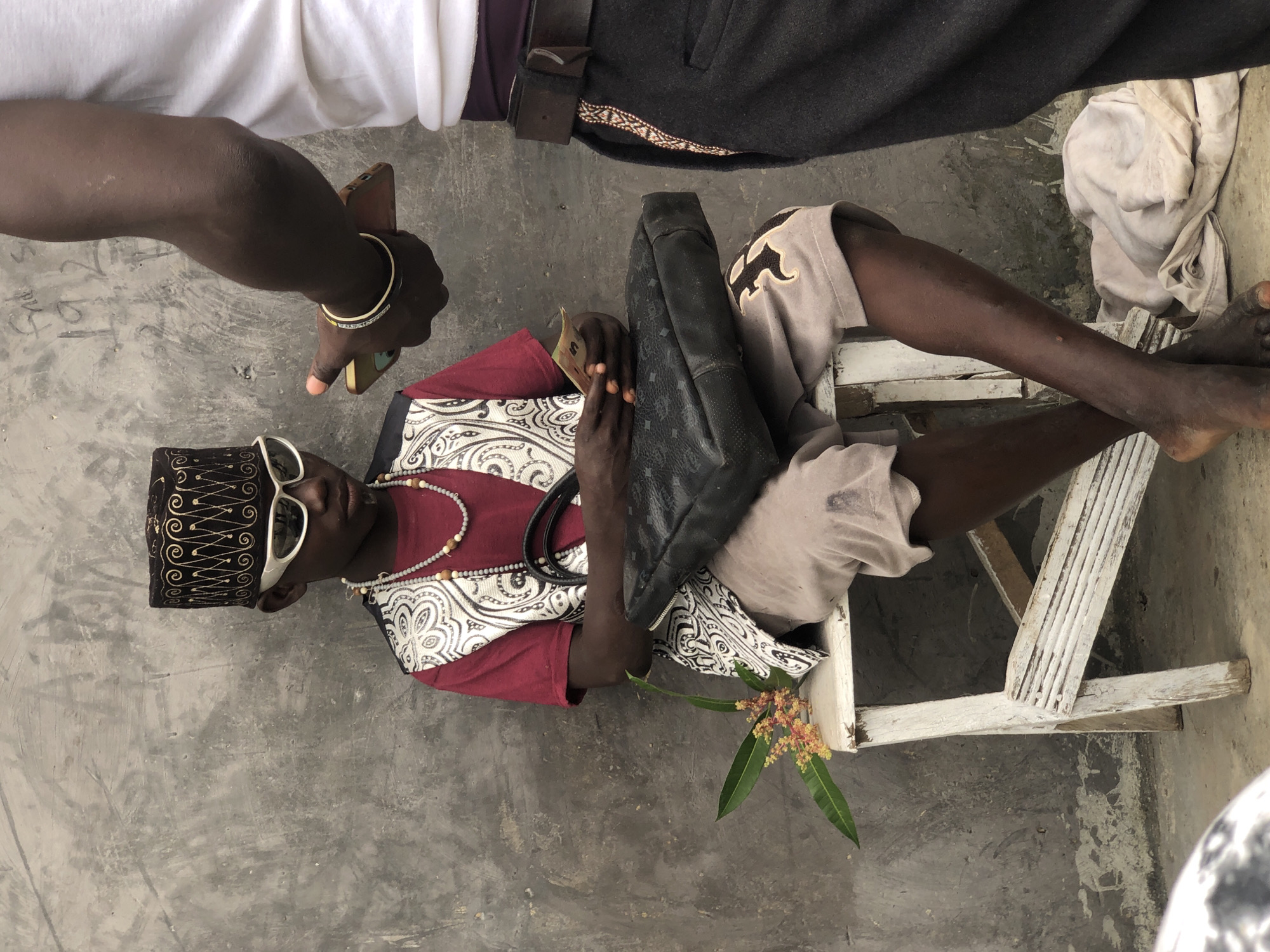 A young Black male, dressed in a maroon shirt, a patterned vest, a traditional cap, and white sunglasses, is seated on a white wooden stool in front of a textured gray wall in Ado, Nigeria. He holds a few banknotes in his hands and a black bag rests on his lap. A small branch with orange and yellow flowers lies on the stool next to him. To the right, another Black person, whose body is partially visible, is holding a smartphone. The environment appears to be an outdoor setting with a rough concrete ground.