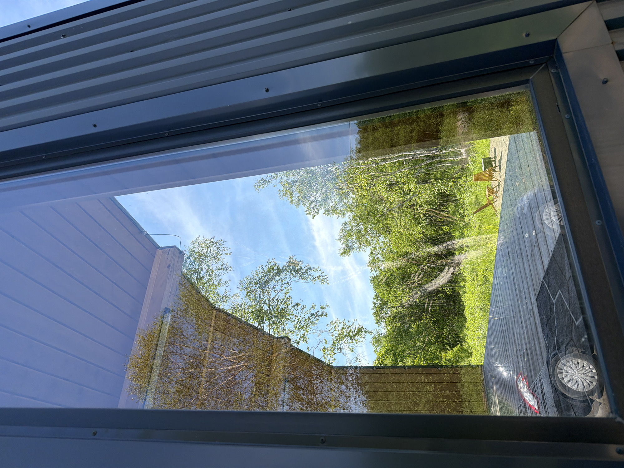 The image depicts a reflection in a large window of a modern structure, likely a house or cabin, in the city of Novy, Russia. The reflection shows a bright, sunny day with a blue sky dotted with white clouds. Lush green trees and foliage are visible, suggesting a natural, possibly wooded environment surrounding the building. In the foreground of the reflection, a dark-colored car is parked on a wooden deck. Two empty chairs and a small table are also present on the deck, indicating an outdoor seating area. The exterior of the building itself is visible on the right side of the image, featuring dark, corrugated metal siding. The window frames are dark gray.