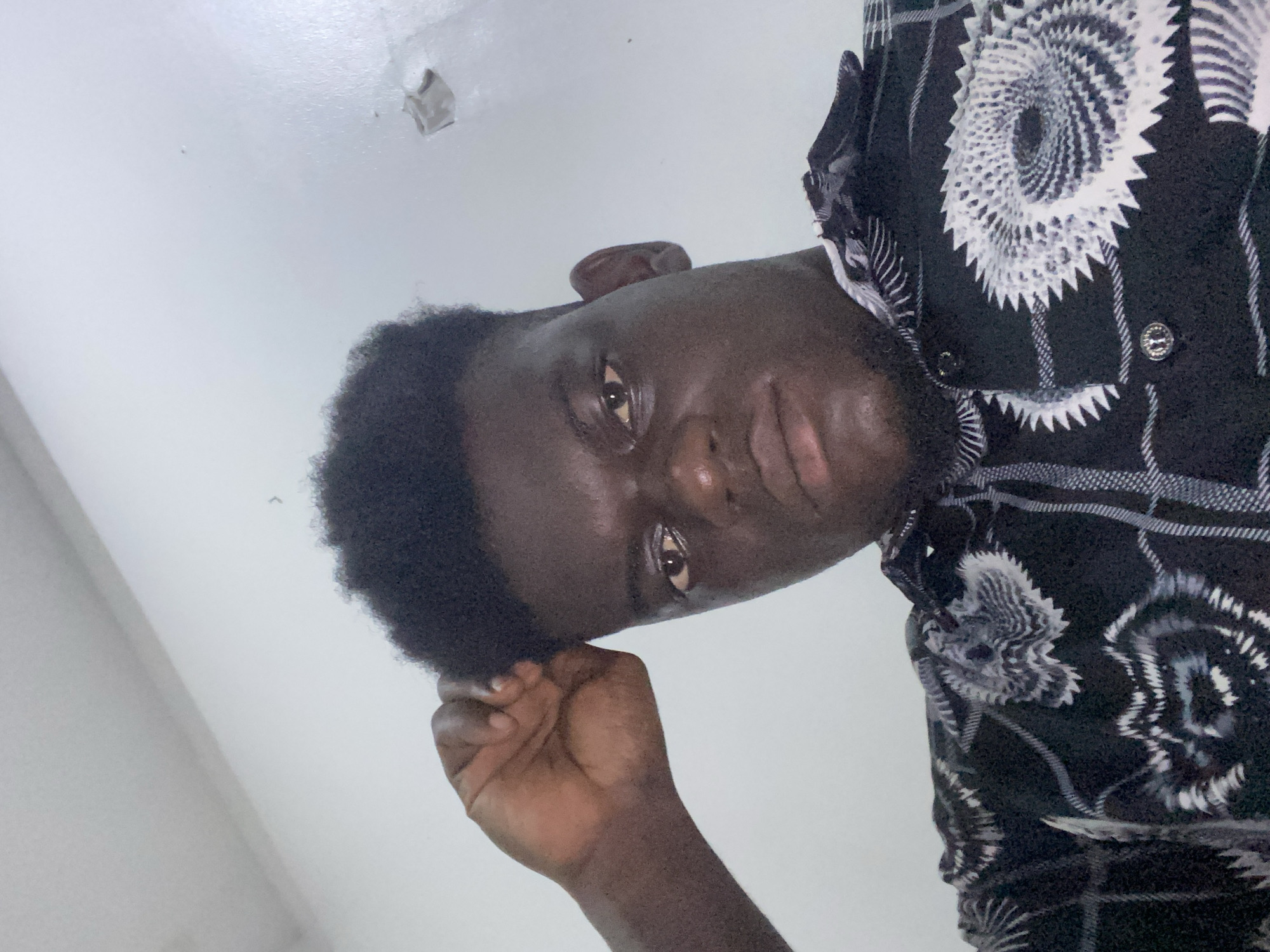 The image displays a young man with dark skin, captured from a low-angle perspective, with his head tilted diagonally as if looking upwards. His gaze is directed towards the camera, conveying a neutral to slightly serious expression. He has short, dark, curly hair and a faint beard. His right hand is visible, resting near his temple or hair on the left side of the frame.
He is wearing a short-sleeved, button-up shirt that is predominantly black, featuring an intricate pattern of white and grey. This pattern includes large, circular, gear-like or spiral motifs with jagged edges, interspersed with finer white lines forming a grid or plaid design. Two metallic buttons are visible on the shirt's front.
The background is a plain, light-colored surface, likely an indoor ceiling or wall, appearing white or off-white. A minor imperfection, possibly peeling paint or a small hole, is noticeable on this surface in the upper right area of the image. The lighting is soft and even, suggesting an indoor setting. No text is visible in the image.