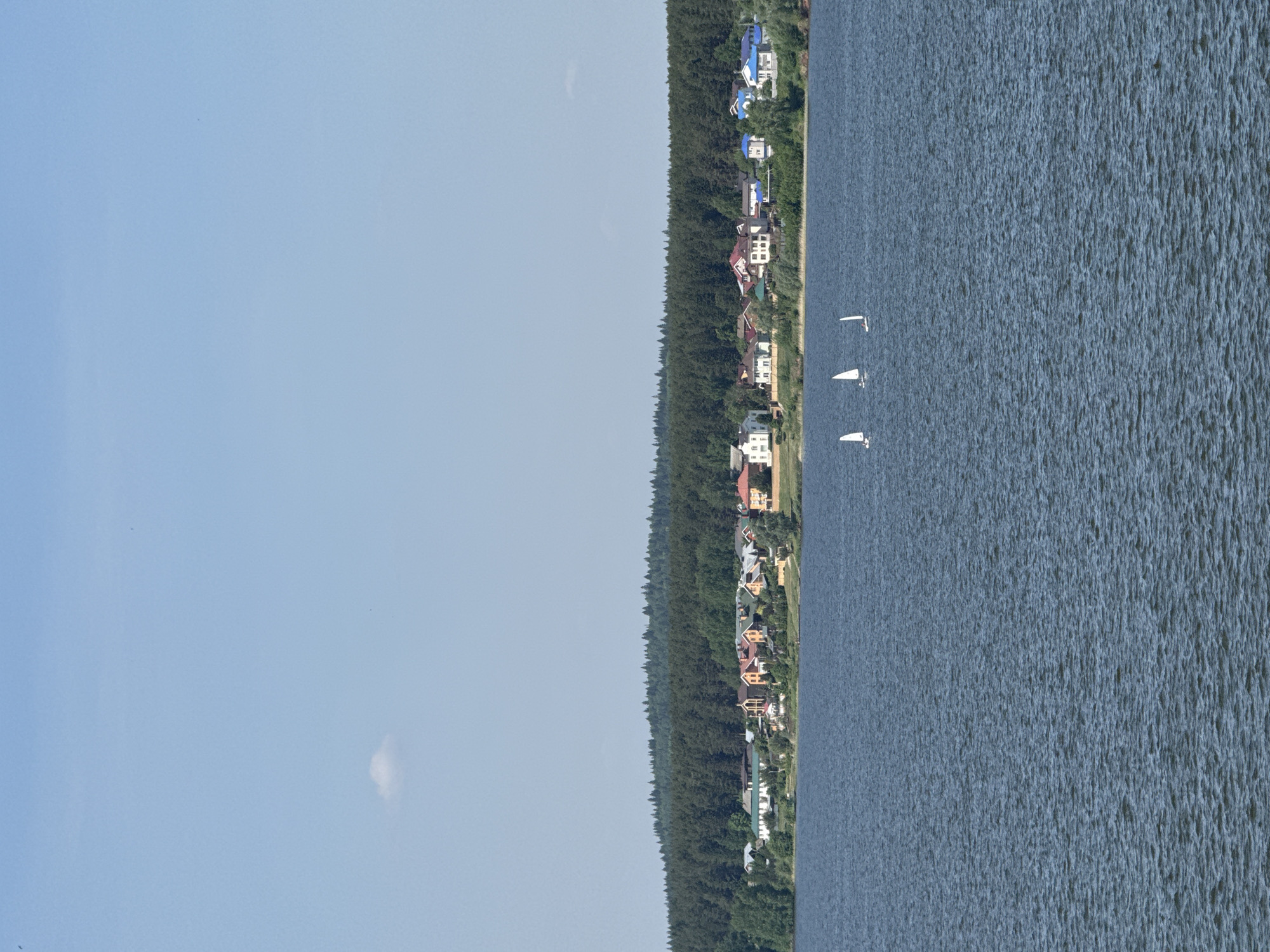 The image depicts a lakeside scene near Votkinsk, Russia. A line of houses borders a dense forest along the water's edge. Three sailboats are visible on the water. The sky is pale blue and mostly clear.