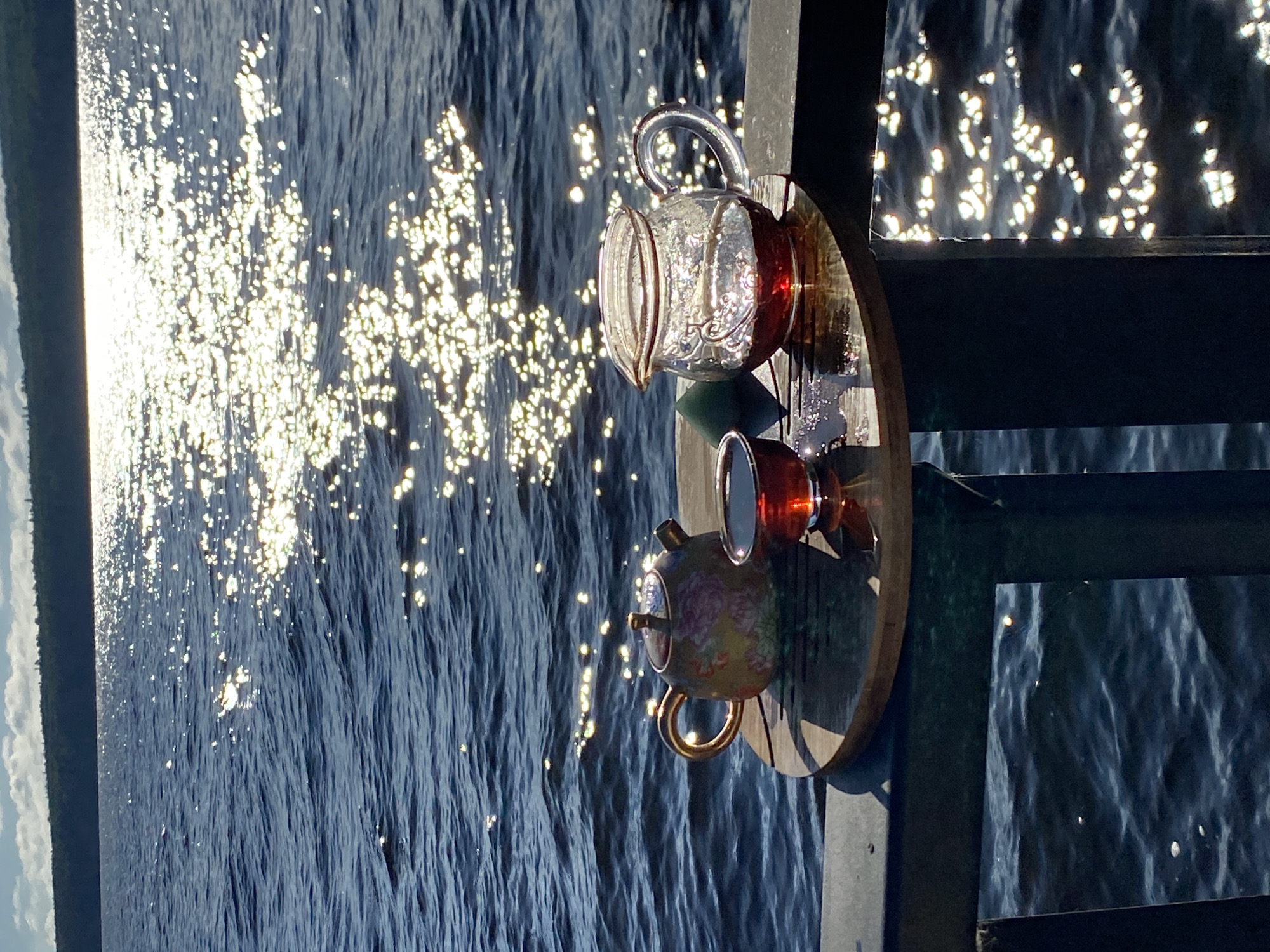 On a wooden board placed on a railing, a tea set is arranged by the water. The set includes a small, ornate teapot with a floral design and a gold handle, a dark red teacup filled with tea, and a clear glass pitcher also containing tea. A small, dark green object, possibly a jade stone, sits next to the pitcher. The board is positioned at the edge of a body of water, which reflects the bright sunlight, creating a dazzling effect of sparkles on the dark blue surface. In the background, a tree-lined horizon is visible under a partly cloudy sky. The overall scene suggests a moment of peaceful contemplation by the water, possibly in Chaikovsky, Russia, a city known for its picturesque natural surroundings.