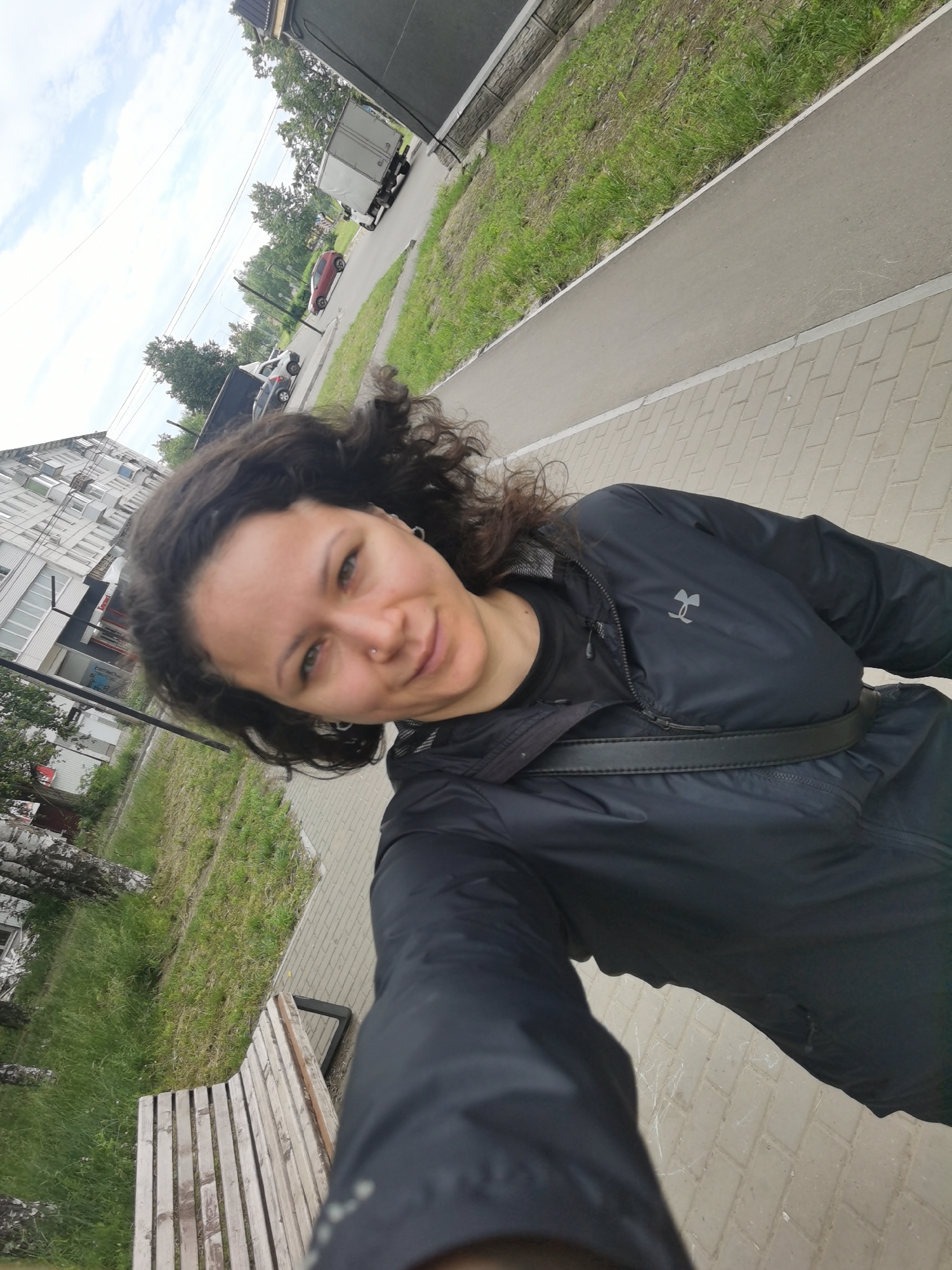A first-person selfie shows a woman with curly dark hair and green eyes, wearing a black Under Armour jacket and a black t-shirt. She has a nose ring and hoop earrings. She is standing on a paved sidewalk next to a grassy area with trees and a bench. In the background, there is a multi-story building, a road with cars and a truck, and a cloudy sky. The image appears to be taken in a city setting, likely in Chaikovsky, Russia, given the architecture and vegetation. The overall scene suggests a casual outdoor activity on a moderately overcast day.