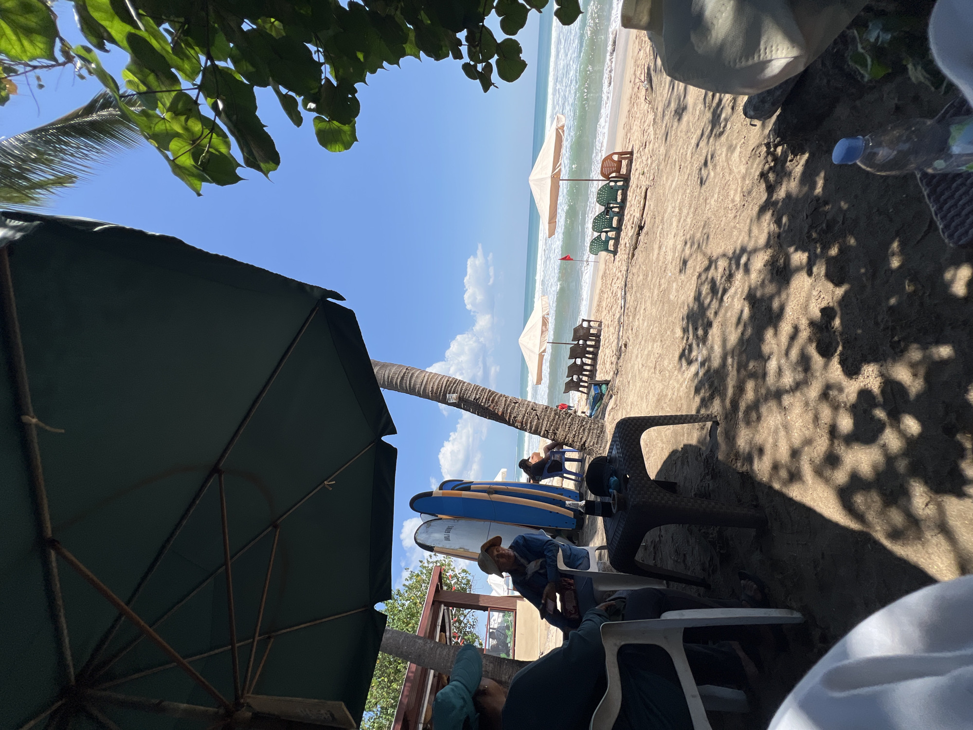 This image depicts a relaxed scene at a beachside cafe in Kuta, Indonesia. The foreground is dominated by a large, dark green umbrella providing shade over a sandy area where two people are seated. To the left, a person wearing a blue shirt and a blue hat is seated in a white plastic chair. In the center, another person with a straw hat and a denim jacket is also seated, holding a smartphone. A small, dark plastic table sits between them, holding a bottle, a hat, and an ashtray.
Behind them, several surfboards are propped up, with two prominent blue surfboards featuring orange trim. A coconut palm tree trunk is visible on the right. The background reveals the beach with a gentle surf and clear blue sky. Two beige beach umbrellas are set up on the sand, with rows of empty green and orange plastic chairs arranged in front of them. A small red flag is planted in the sand near one of the umbrellas. The overall atmosphere suggests a peaceful day at the beach, likely a popular spot for surfers and tourists.