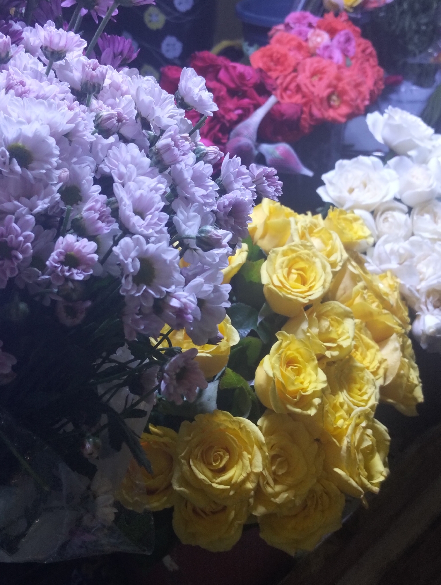The image shows a close-up of several bunches of flowers. From left to right, there's a bouquet of light purple chrysanthemums, followed by yellow roses, and white roses. In the background, there are two more bunches of flowers, one red and the other deep red. The photo appears to be taken in a shop or flower market, given the variety and density of the floral displays. The precise location in Dhaka, Bangladesh cannot be determined from the visual information provided. There are no people visible in the frame.
