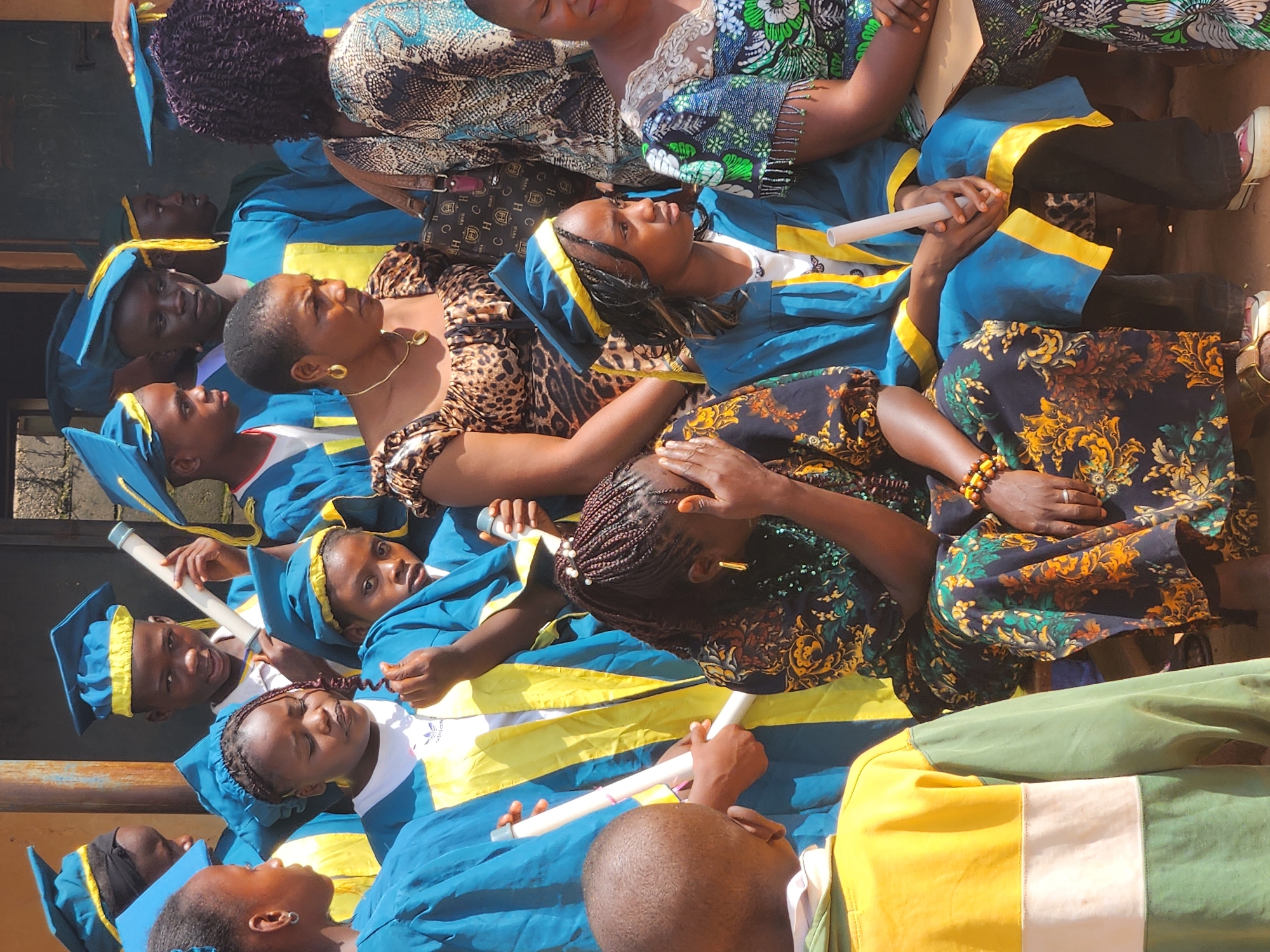 The image depicts a group of individuals, primarily children and young adults, dressed in blue and yellow graduation gowns and caps. They appear to be participating in a graduation ceremony or a similar event. In the foreground, a young girl with braided hair, wearing a graduation gown and a dark patterned dress, has her hand covering her face, seemingly in distress. To her left, a young boy in a yellow and green shirt is partially visible from the back. Behind them, other children in graduation attire are looking in various directions. To the right, two women are also present, one in a leopard print top and the other in a patterned dress. The background suggests an outdoor setting, possibly near a building with a textured wall and a wooden structure. The lighting indicates it is daytime. The scene is set in Sabon Tasha, Nigeria, a location known for its vibrant community and educational institutions.
