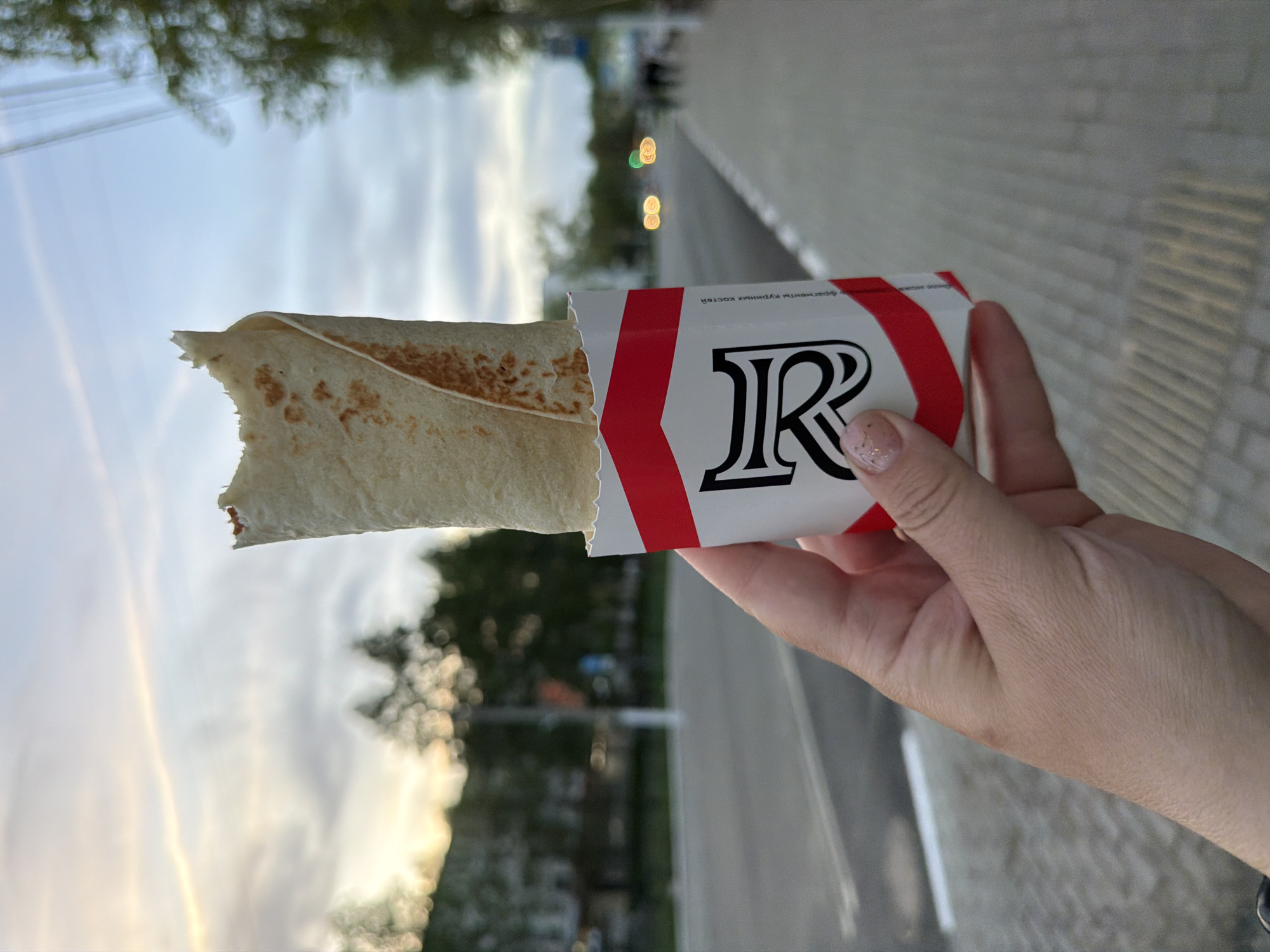 A hand holds a partially consumed shawarma (a type of wrap or sandwich) in a white cardboard wrapper adorned with red chevron stripes and a stylized black letter "R," outdoors. The shawarma's contents are visible at the open end of the wrapper, the thin tortilla revealing the meat and other filling. The person holding the shawarma has a light complexion, visible skin texture, and manicured nails with a glittery polish. The background is blurred, but suggests a city street in Chaikovsky, Russia with visible paved surfaces (possibly a walkway or sidewalk) and glimpses of trees and sky. Street lights are vaguely visible in the background. The image is captured at an angle, possibly indicating that the picture was taken while walking.