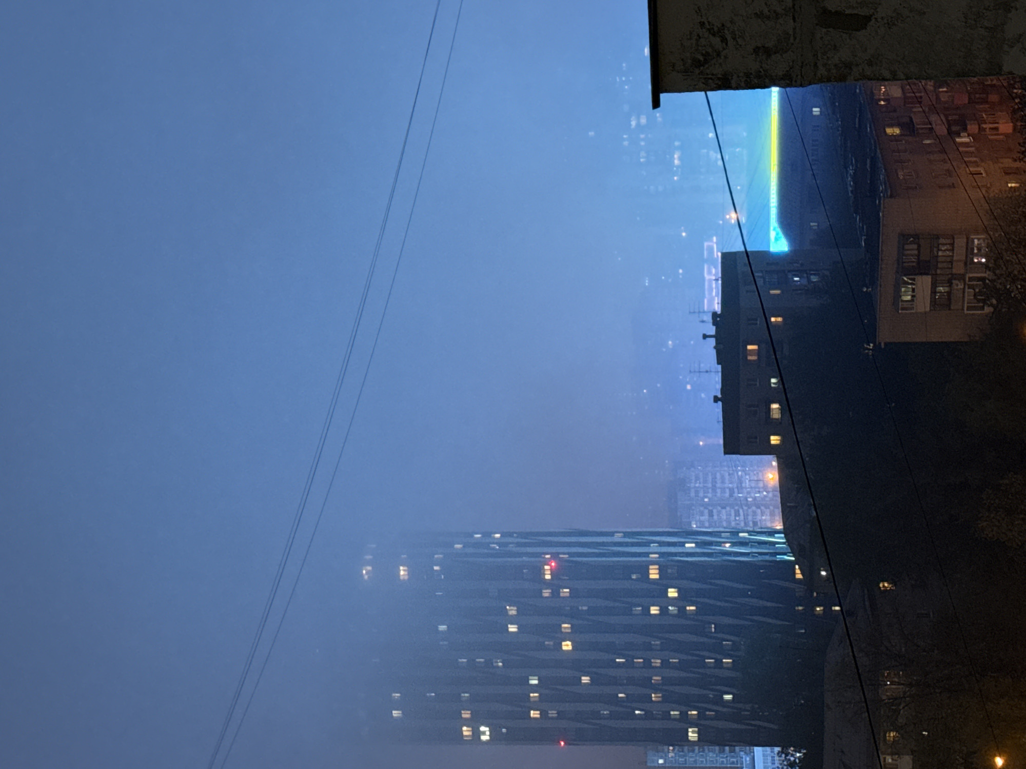 The photograph captures an urban nightscape in Yekaterinburg, Russia, viewed from an elevated vantage point, likely a window. The scene is dominated by a dense fog or mist, partially obscuring buildings and streetlights. Power lines diagonally traverse the upper-left portion of the frame.
In the foreground, portions of two buildings border the right edge of the image. These structures appear older and contain a number of lit and unlit windows.
Beyond these buildings, a cluster of illuminated high-rise apartments pierces through the fog. Their windows display a mixture of light patterns, indicating varying occupancy. A beam of blue and yellow light stretches into the skyline.