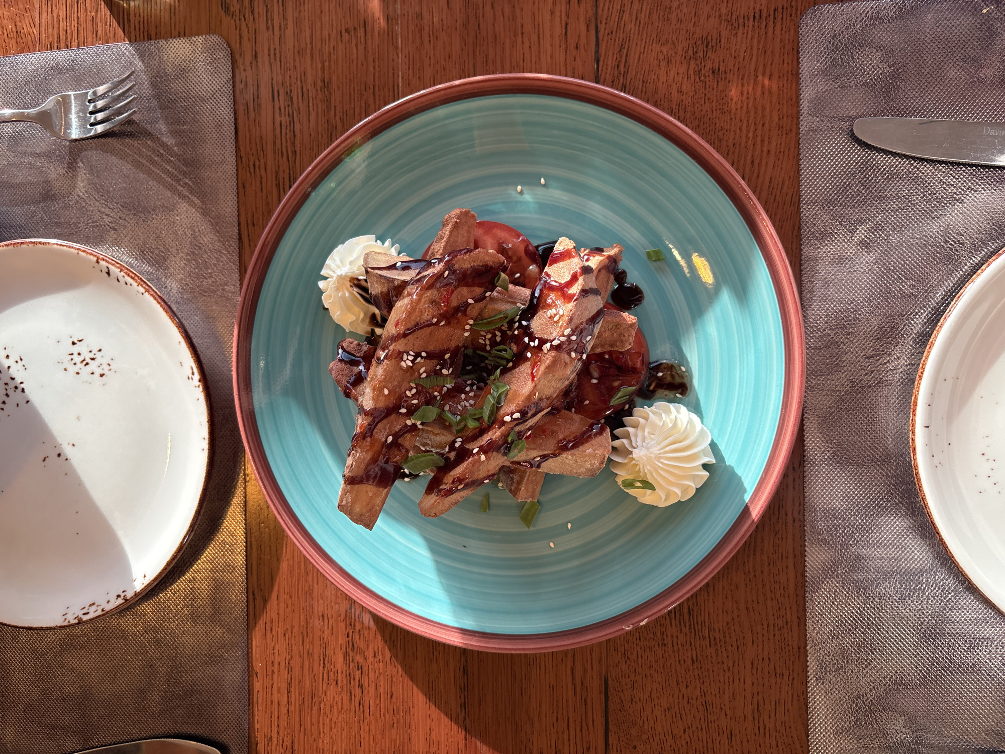 A overhead, close-up shot shows a turquoise plate filled with french fries drizzled with a dark sauce and sprinkled with sesame seeds and chopped green onions. Two dollops of whipped cream are placed on the side of the plate. The plate is on a dark wooden table with a brown placemat and a white plate in the background. The setting appears to be a restaurant in Saint Petersburg, Russia, suggested by the presence of traditional Russian nesting dolls in the background and the overall aesthetic often found in St. Petersburg eateries. Sunlight is streaming in from the left, casting a warm glow on the scene.