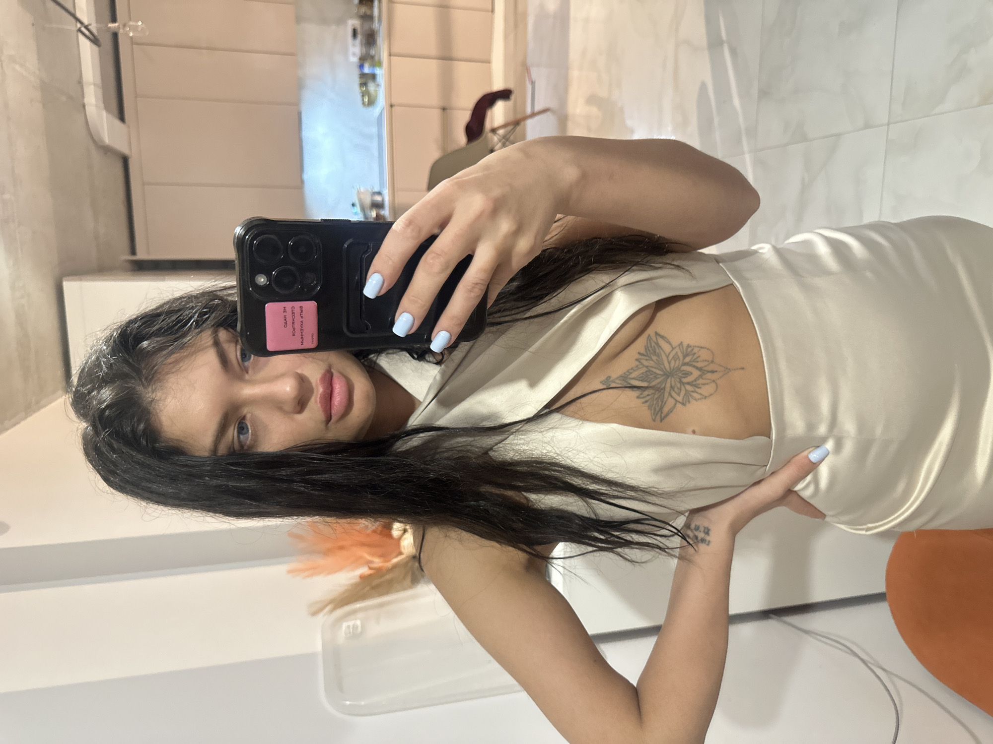 The image is a mirror selfie featuring a young woman in an indoor setting, identified as Khimki, Russia. The woman has long, dark, wavy hair, light-colored eyes, and full pink lips. She is wearing a light-colored, possibly cream or white, satiny top or dress with a deep V-neckline. Her fingernails are painted a light blue.
A prominent tattoo with an intricate floral or mandala design is visible on her upper chest/sternum area. Another tattoo, featuring numbers and symbols (partially readable as "10.09.19.XX"), is present on her left wrist.
She holds a black smartphone, likely an iPhone, in her right hand to take the picture. The phone is encased in a black cover, and a pink rectangular sticker is adhered to its back. Due to the reflection in the mirror, the text on this sticker appears reversed, reading "СЛИШКОМ УМНА ЧТОБЫ БЫТЬ ОДНОЙ" (SLISHKOM UMNA CHTOBY BYT' ODNOY), which translates to "Too smart to be alone." Below this, another word, "ОФИГЕТЬ" (OFIGET'), a colloquial Russian exclamation, is partially visible.
The background, seen through the reflection, depicts a modern interior space. It features light-colored walls, some with horizontal paneling, and a light-toned tiled floor. Blurred elements of furniture, including a chair and shelves with bottles, are subtly visible in the distance. An orange-colored object, possibly a cushion or part of a furniture piece, is present in the lower right portion of the reflection. The woman maintains a neutral expression, looking directly at the phone's camera. The lighting suggests an indoor environment.