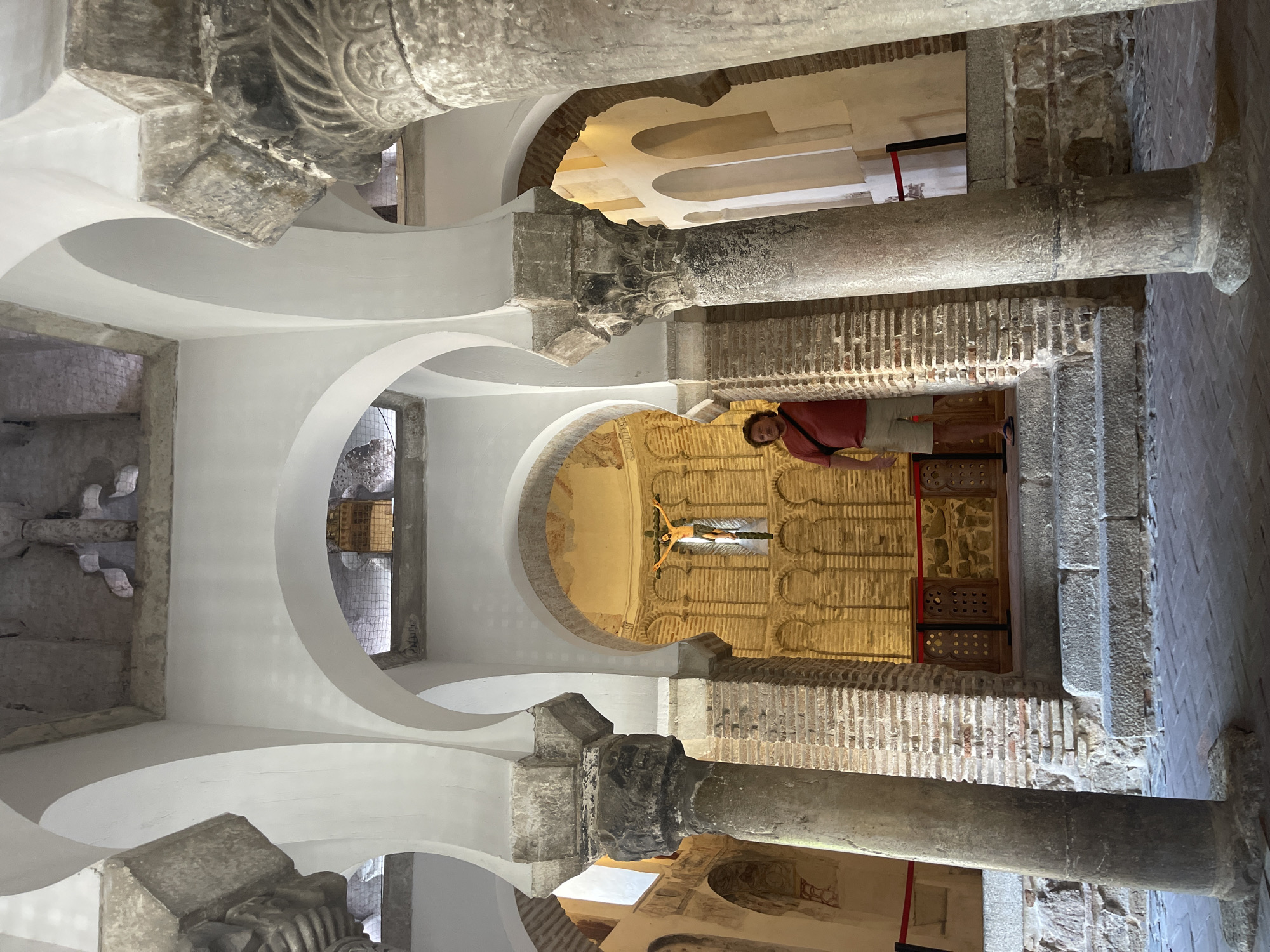 A man stands in a courtyard with Moorish arches and columns, facing a wall with a crucifix. The courtyard is constructed of brick and stone, with patterned tile work on the floor. The man is wearing a red shirt and beige shorts, with a black bag slung over his shoulder. He is standing on a set of stone steps, looking towards the crucifix. The scene is illuminated by natural light streaming in from an unseen source. This image appears to be taken inside the Synagogue of Santa María la Blanca, a historic building in Toledo, Spain, known for its unique blend of Mudejar and Gothic architecture.