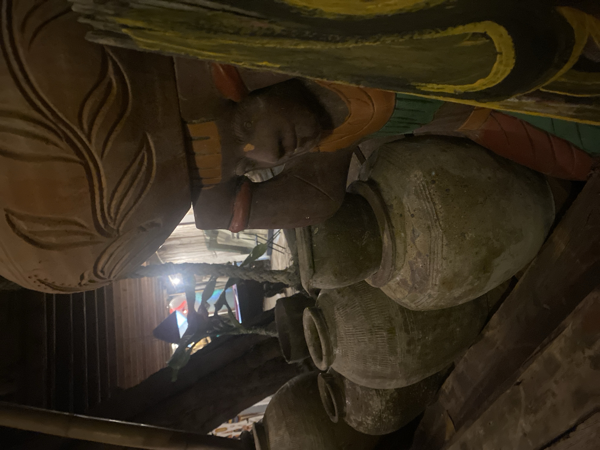 The image shows a close-up, upward-angle view of carved wooden figures and a stack of earthen pots in Baan Don Sai, Thailand. The figures appear to be part of a larger structure or sculpture, with one figure depicting a human face and body adorned with colored accents (yellow, green, red). The pots are stacked vertically, their surfaces textured and aged. A rope or vine is visible in the background, along with glimpses of an interior space, including wooden beams, corrugated metal, and colored lighting.
