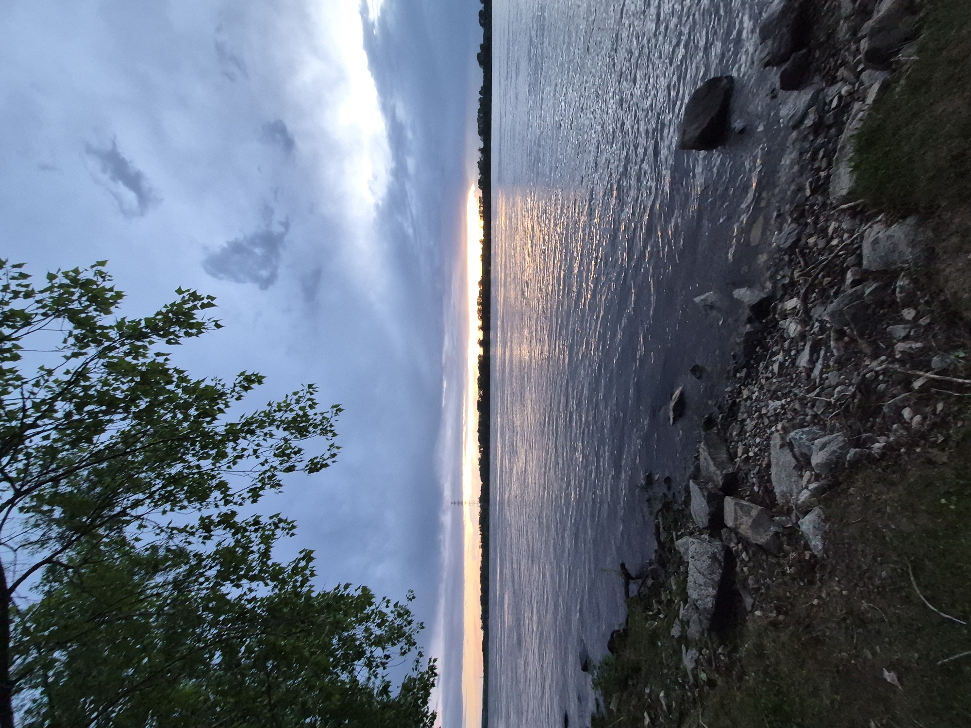 The image depicts a landscape view in Chaikovsky, Russia, possibly near a riverbank or lake during sunset. A body of water occupies the right half of the frame with the fading sunlight reflecting across its surface. The shoreline is rocky, with various-sized stones and some vegetation along the edge. On the left, a tree with green foliage frames the scene. The sky overhead is cloudy, with varying shades of blue and gray, indicating the approach of twilight. In the center horizon, a thin line of orange and yellow light marks the setting sun. Transmission towers are visible along the sunset horizon.