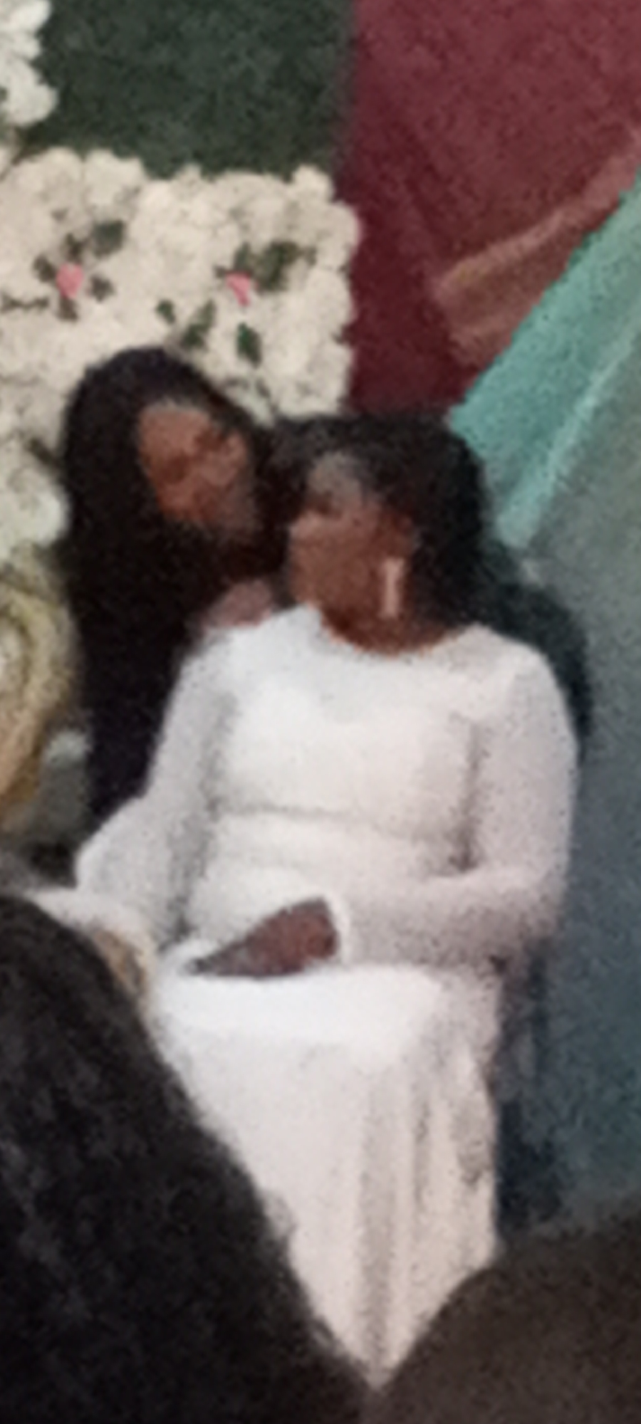 Two women are in the image. One woman wears a white dress and sits down. The other woman, with dark hair, stands behind her. They appear to be interacting or looking at each other. There are white flowers in the background. The scene appears to be indoors. It is not possible to confirm if the image was taken in Kakuri Gwari, Nigeria.