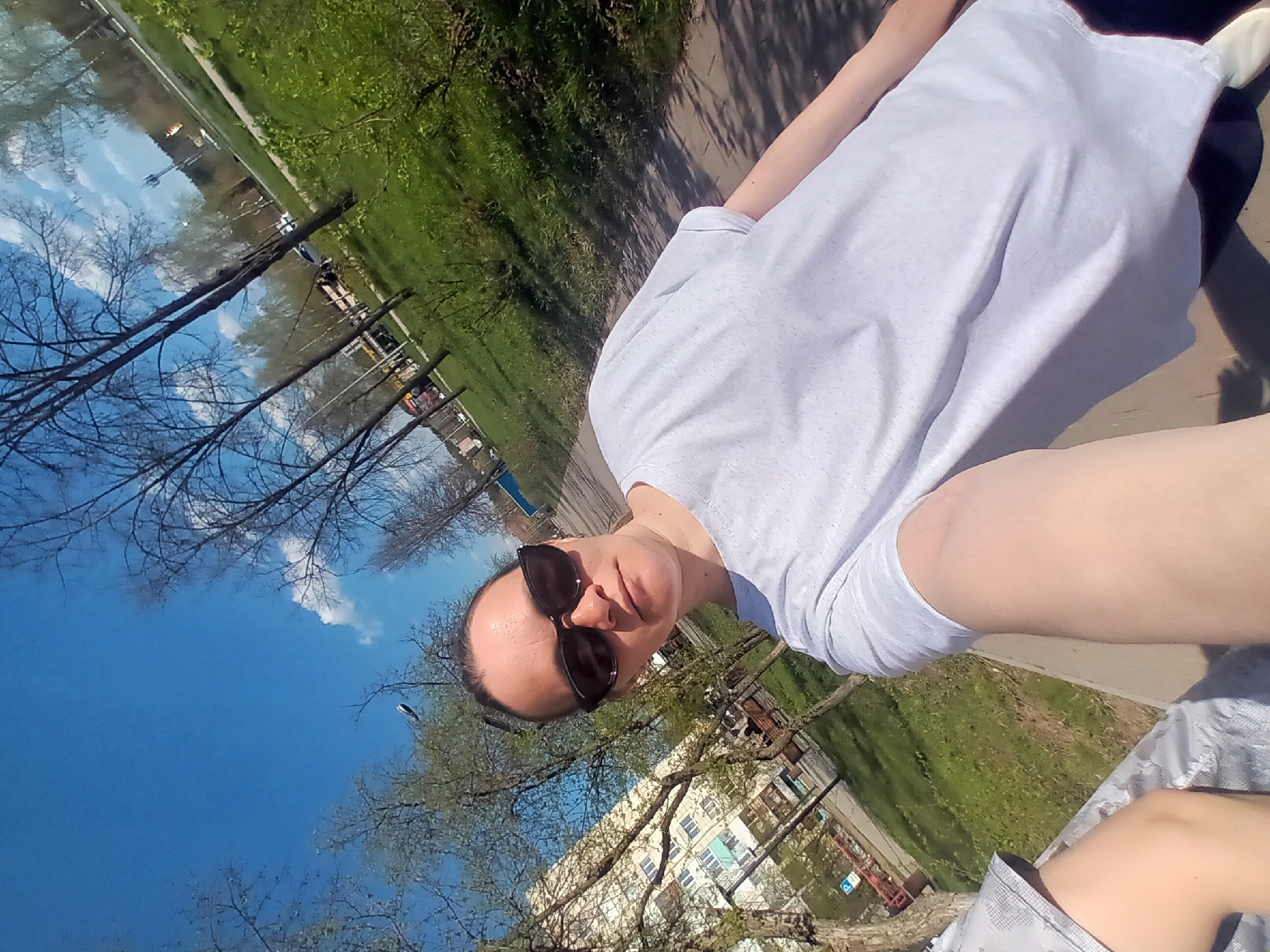 The image shows a person with sunglasses wearing a white shirt, standing on a walkway in Chaikovsky, Russia. The background includes trees, grass, a lake, and buildings. The sky is blue.