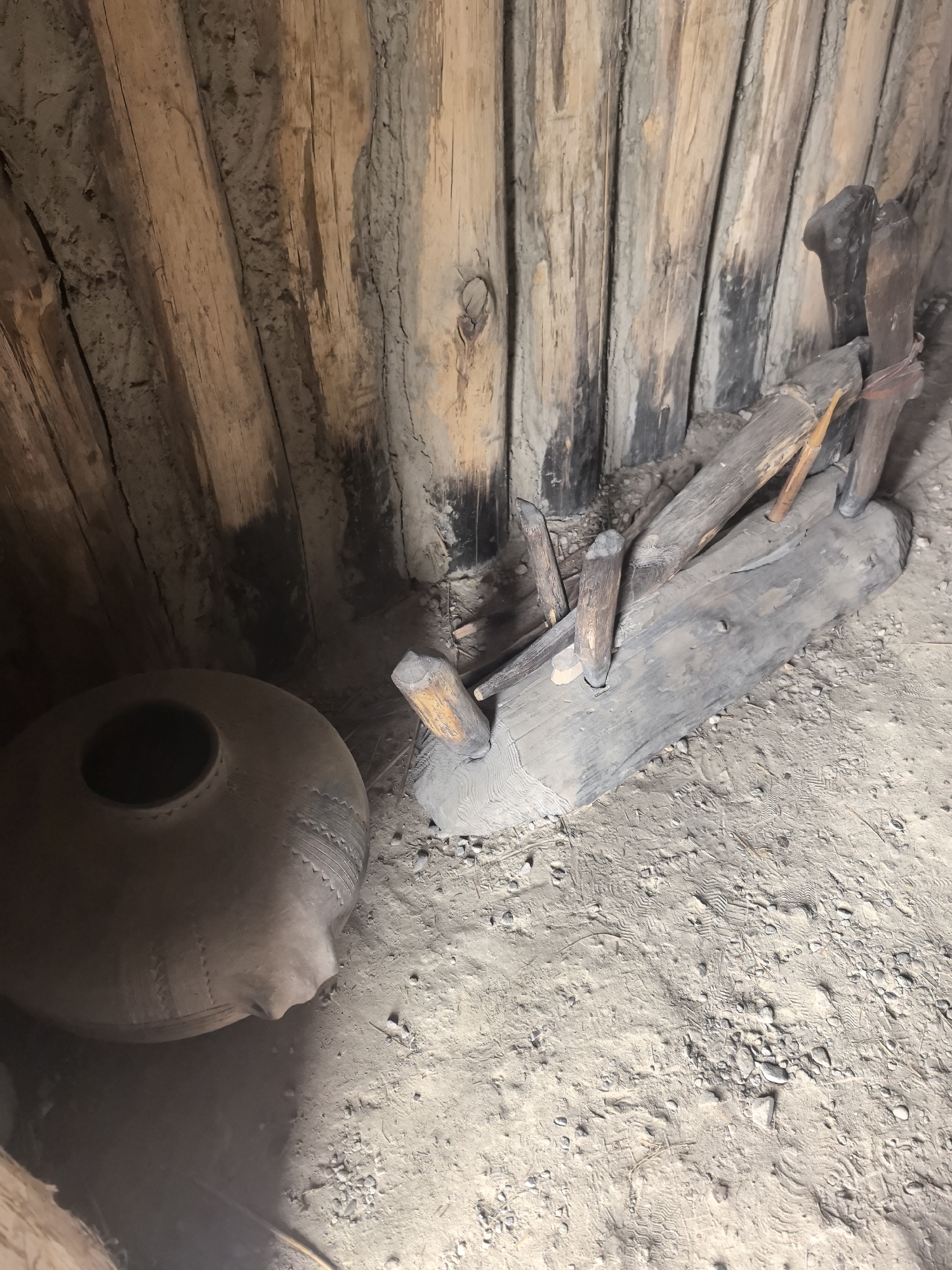 A wide, high-angle shot shows the interior of a rustic, wooden structure with a dirt floor. The walls are constructed from vertical, rough-hewn logs, some of which appear charred. On the left side of the frame sits a large, round, clay pot with a wide opening and decorative bands. To the right of the pot, a large, weathered wooden beam is positioned horizontally on the dirt floor, with several smaller wooden stakes or pieces of wood protruding from it and embedded in the ground. The ground is covered in sandy soil, scattered with small stones and debris, and appears to have several footprints. The lighting is dim and appears to be natural light filtering into the structure. This scene is located within the Камянюцкі сельскі Савет, Belarus.