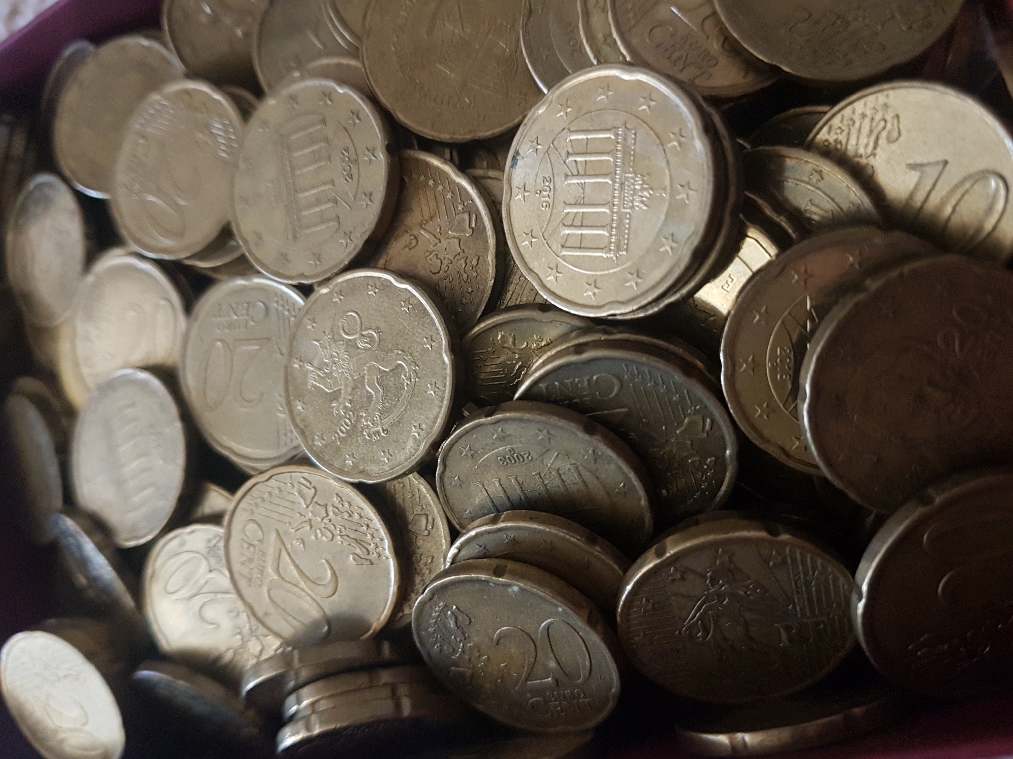 The image depicts a close-up of a collection of Euro coins. The coins are predominantly 20 Euro cent and 10 Euro cent pieces. The coins feature different designs and dates, including images of buildings and national symbols from various European countries. The coins are piled together and appear to be stored in a container or tray. The lighting is somewhat dim, giving the coins a slightly muted, golden tone. There is no indication of Saint Petersburg, Russia, or any people in the image.