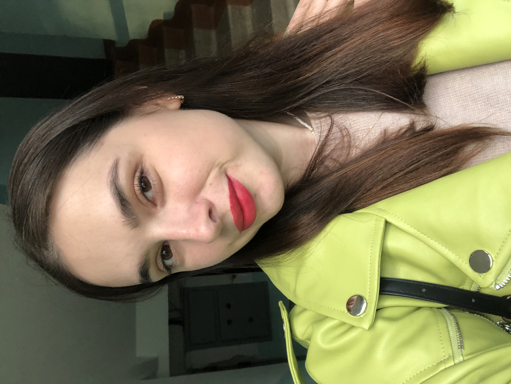 A young woman with long, dark brown hair and brown eyes is captured in a close-up selfie. She is wearing a bright, lime green leather jacket over a light pink textured sweater. Her lips are painted with a bold red lipstick, and she has a slight smile. A thin necklace with small white beads is visible around her neck. She is also wearing small hoop earrings with clear embellishments. Her right hand is resting on her shoulder, and her left shoulder is partially visible. The background is out of focus, but appears to be an indoor setting with a stairway on the right and a dark panel on the left. The lighting is soft and even. The location is the city of Chaikovsky, Russia.