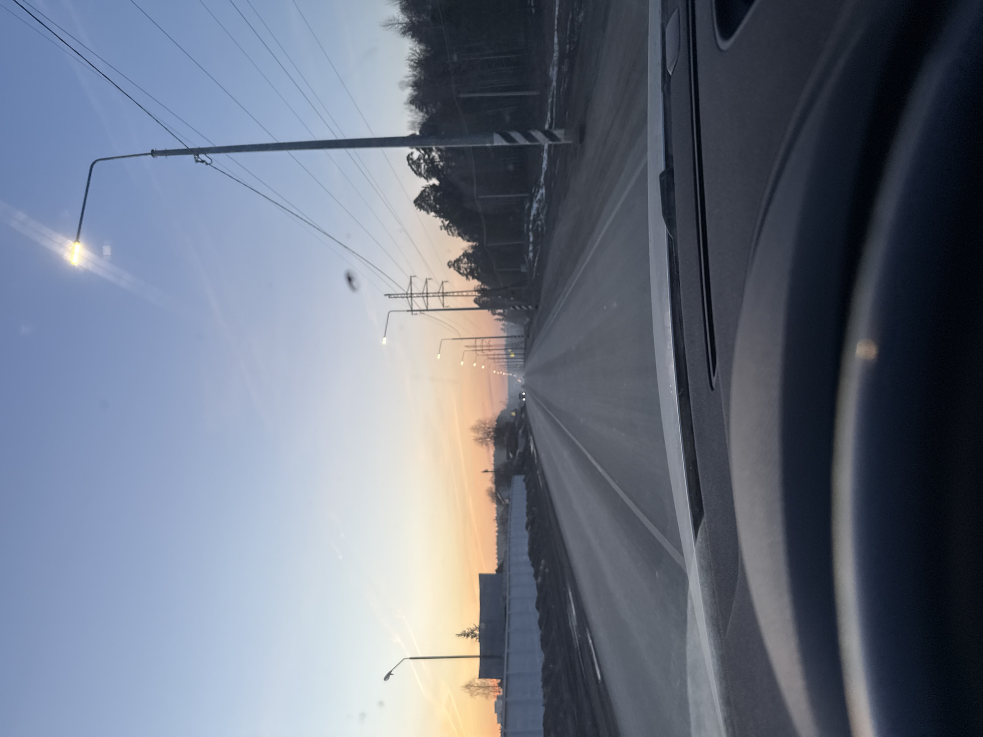 The image depicts a road, likely in the city of Chaikovsky, Russia, as suggested by the environment. The view is from inside a vehicle, with part of the car interior visible. The road is straight, with streetlights lining the sides. Power lines run parallel to the road. The sky is a gradient of colors, suggesting either sunrise or sunset. On one side of the road, there is a guardrail and a glimpse of trees. The presence of trees and their bare appearance implies it is likely either winter or late autumn. No people are visible.