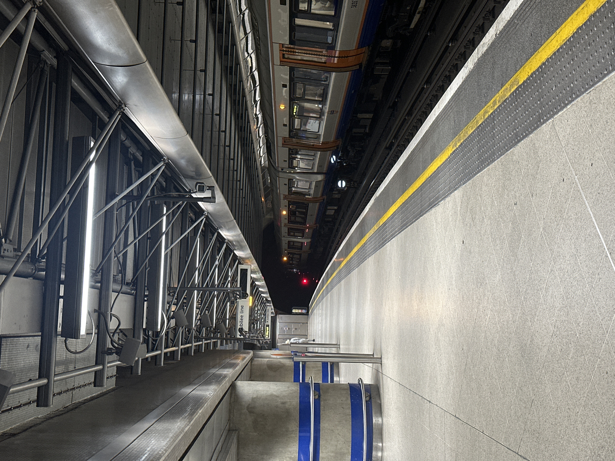 The image depicts the interior of a London Underground station, specifically on the Jubilee Line platform. A train is visible on the right side of the frame, aligned with the platform. The platform edge is marked with a yellow safety line. On the left, a metallic structure supports the platform and possibly overhead lines. Stairs with blue markings lead down from the platform level. The overall impression is one of a modern, functional, and somewhat industrial underground transportation hub.