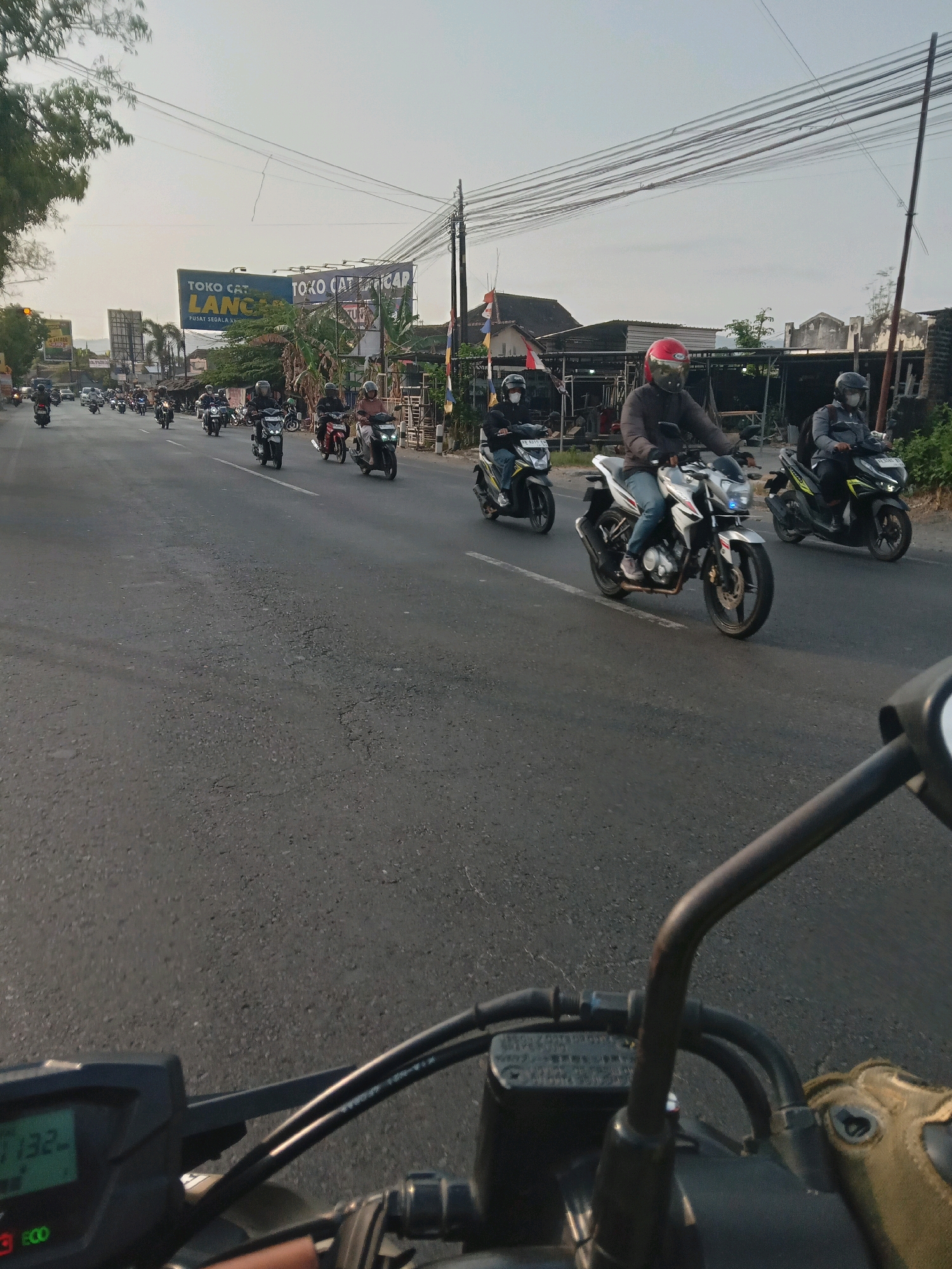 This image depicts a busy street scene in Berbah, Indonesia, likely during daylight hours, as indicated by the bright sky and shadows. The perspective suggests the photograph is taken from the rider of a motorcycle, with the handlebars and dashboard of the motorcycle visible in the foreground.
The main focus of the image is a procession of motorcycles traveling along a paved road. Numerous individuals are riding motorcycles, most wearing helmets for safety. The motorcycles vary in style, including scooters and what appear to be more standard motorcycle models. Some riders are alone, while others may be carrying passengers, though this is not always discernible. The motorcycles are moving in the same direction, suggesting a convoy or a common route.
Along the sides of the road, there are various structures. On the left, there are trees and what appear to be buildings or shops. A prominent billboard is visible, advertising "TOKO CAT LANCAR" (Lancar Paint Store) with text below indicating "PUSAT SEGALA KEBUTUHAN" (Center for All Needs). Further down the road, there are more buildings, some with awnings, and utility poles with a complex network of wires strung between them. The road itself is asphalt and shows signs of wear, with some cracks visible. The overall environment suggests an urban or semi-urban setting. The sky is clear with a slight haze. The direction of travel and the presence of traffic indicate typical daily activity on a road in Berbah.