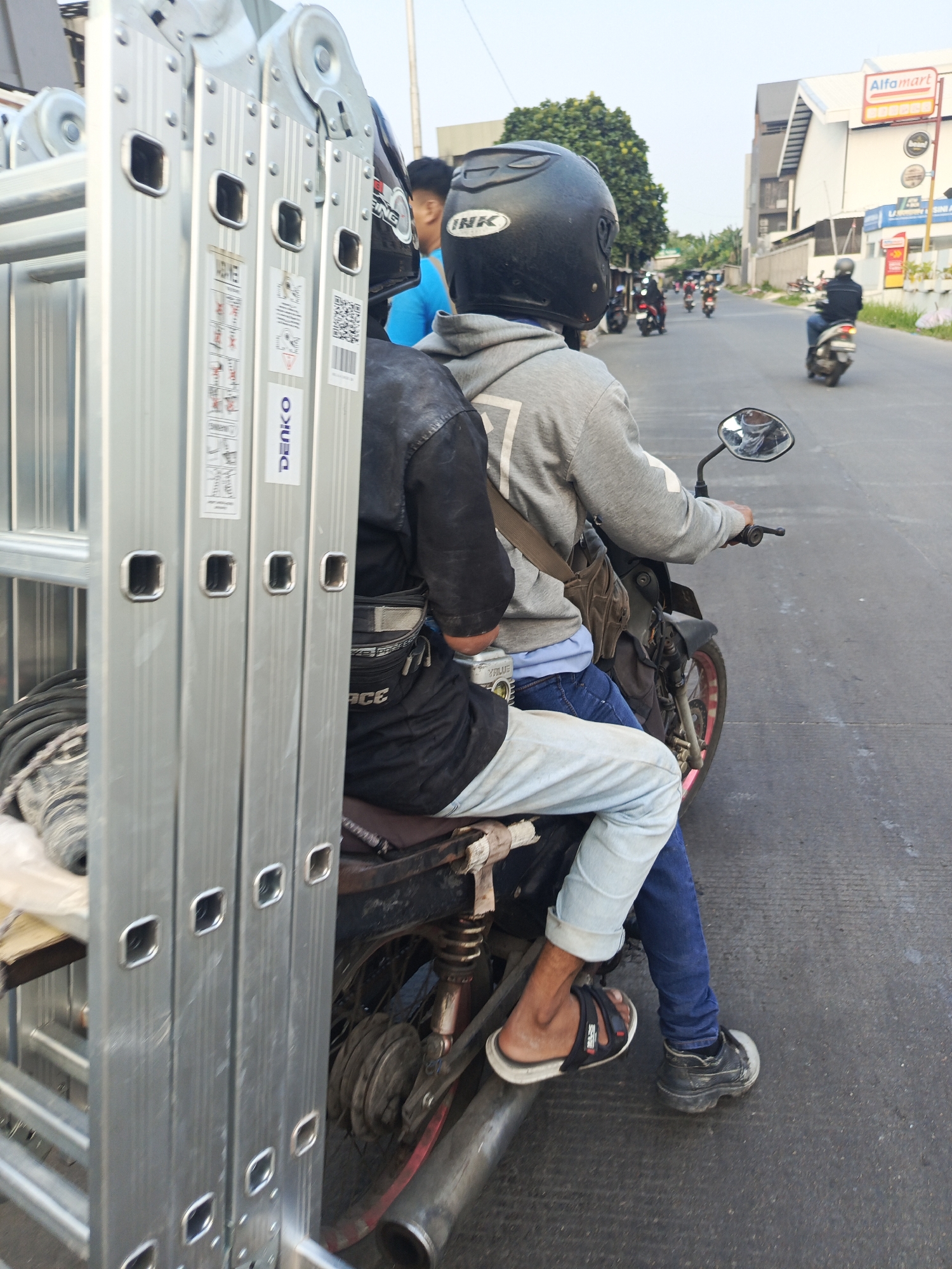 Two individuals are seated on a motorcycle traveling down a paved road in Tangerang, Indonesia. The person in the front is wearing a black helmet and a grey hoodie, with a brown crossbody bag slung over their shoulder. The person behind them is wearing a black jacket and light blue jeans, and they have a black waist bag. Both individuals are wearing footwear appropriate for riding a motorcycle. The motorcycle is carrying a large, silver, multi-section ladder which is positioned vertically along the left side of the vehicle. The ladder is secured to the motorcycle and extends upwards, obscuring part of the rider's view. In the background, there are buildings, trees, and other motorcycles on the road. One of the buildings in the background has a red and white Alfamart sign. The road appears to be a commercial or mixed-use area. The lighting suggests it is daytime.