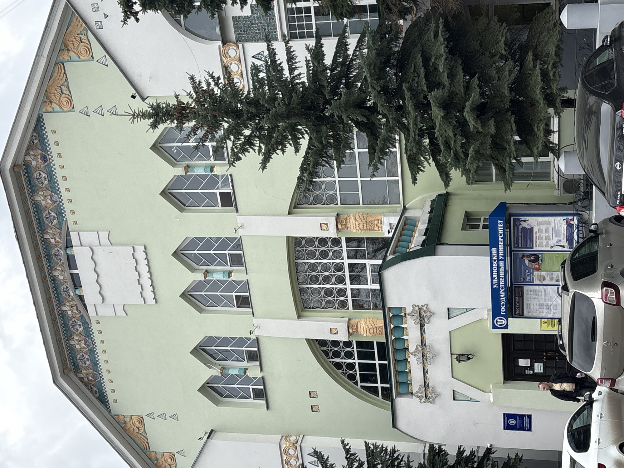 This image depicts the facade of the Ulyanovsk State University building in Ulyanovsk, Russia, on an overcast day. The architecture is distinctive, featuring sections of light green and white. Large evergreen trees partially obscure the building's central portion and the area in front.
The building is richly decorated. Near the roofline on the left, gold-colored, stylized winged motifs are visible, bordered by a band of patterned blue, white, and brown tiles. Small white triangular designs are scattered on the light green wall. Windows vary in shape; some are large arched windows with intricate circular pane patterns, while others are rectangular with geometric, often trapezoidal, divisions. White relief decorations with scalloped and stepped patterns further embellish the light green facade. Ornate white metalwork adorns the white lower section, which also features a gold-colored bust integrated into a structural element.
The main entrance, a dark double door, is centrally located on the lower white section. Above it, a prominent blue sign displays "УЛЬЯНОВСКИЙ ГОСУДАРСТВЕННЫЙ УНИВЕРСИТЕТ" (Ulyanovsk State University) with a circular "УГ" logo, and the word "ПОСТУПАЙ" (Apply/Enroll). A smaller blue sign with the "УГ" logo is positioned below. An information board with various posters stands next to the entrance.
Several cars are parked in front of the building, including a dark Nissan Teana sedan and a silver-colored vehicle. One man, wearing a light-colored top and dark jacket, is visible standing near the entrance, partially obscured by a car. The scene suggests a typical daytime activity at a university campus.
