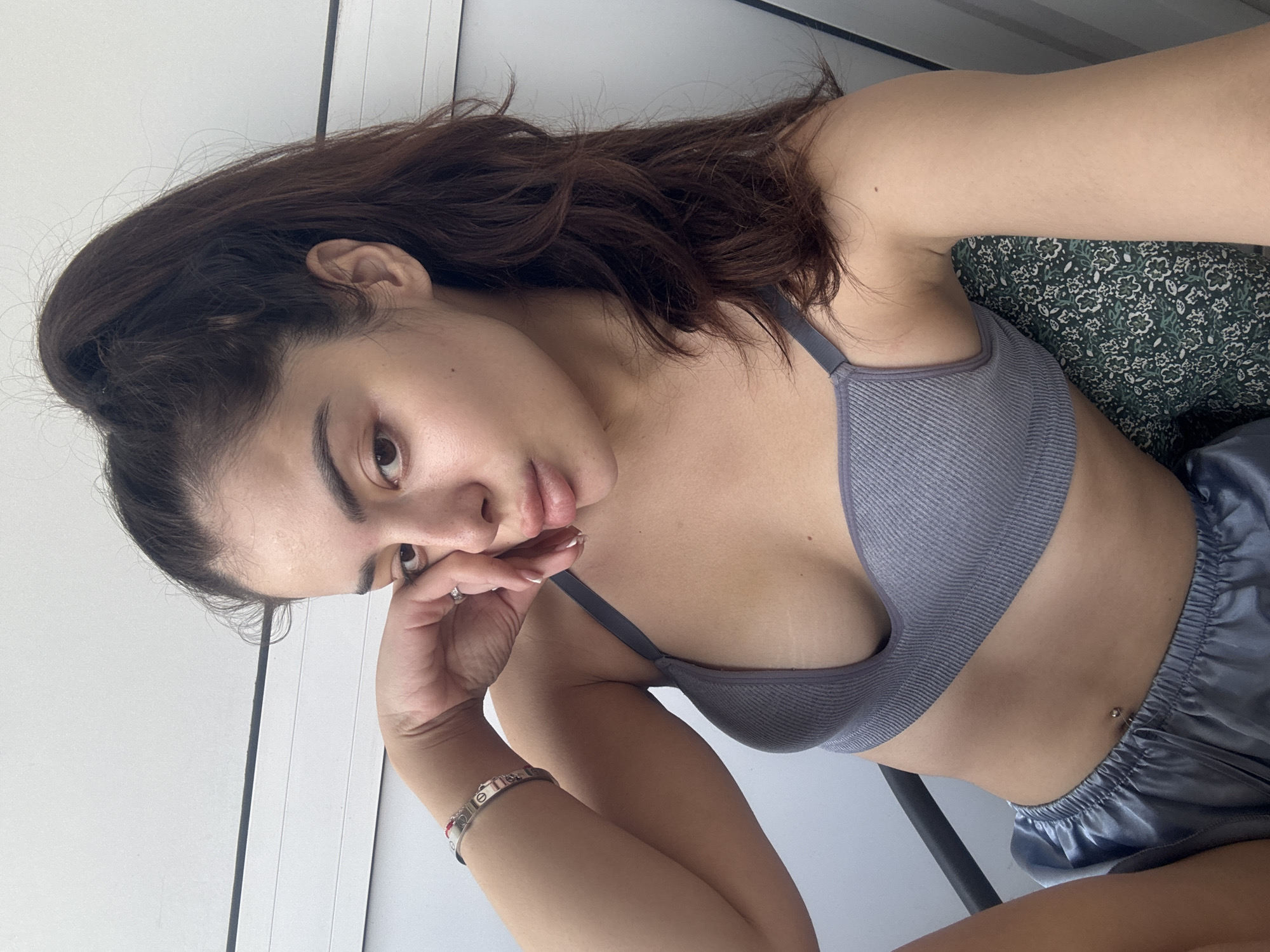A young woman is depicted indoors, possibly in Plovdiv, Bulgaria, posing for a photo while reclining or lying down, with the image rotated 90 degrees clockwise. She has dark brown hair and is wearing a grey ribbed bralette and shiny grey-blue satin-like shorts. Her left hand is resting near her chin, and she looks towards the camera with a relaxed, slightly pensive expression.
Notable details include a silver bracelet on her left wrist, which features the text "CARTIER" and resembles a Cartier Love bracelet, along with a ring on her left ring finger. She also has a belly button piercing visible. The background shows a light-colored wall or door frame and a dark green fabric with a floral or abstract pattern, likely a cushion or part of a chair. The lighting suggests it is daytime.