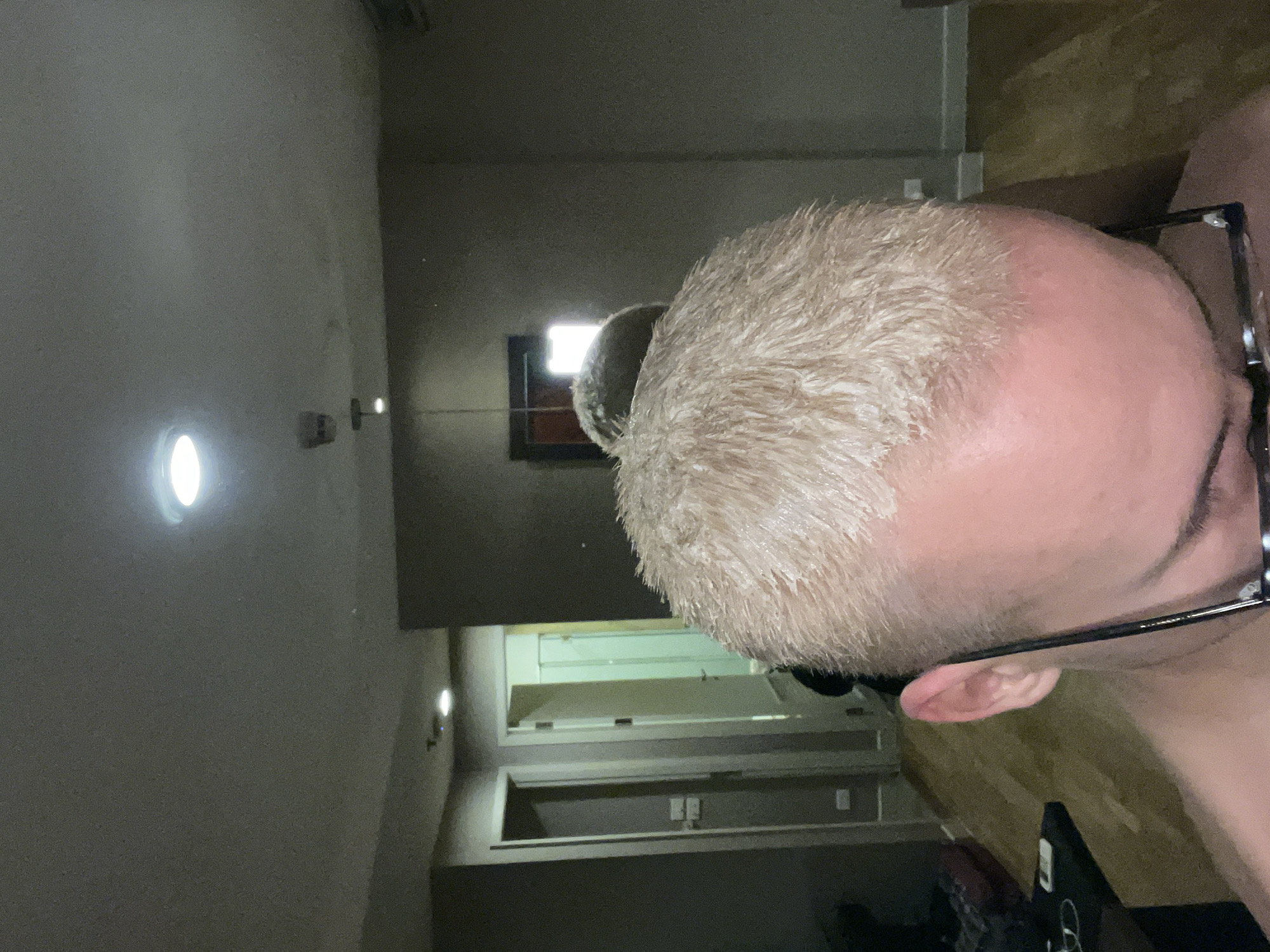 This media file captures a close-up, overhead view of a person's head in an indoor setting, likely in Bangkok, Thailand. The subject has very short, light-colored hair, possibly bleached blonde, which appears slightly damp or textured. The scalp is visible on the upper forehead and crown, exhibiting a light pinkish tone. The person is wearing black-framed glasses, with one arm of the frame extending over their ear.
The background depicts a dimly lit room or hallway. Several artificial light sources illuminate the space, including a prominent circular wall-mounted light on the left and another bright, rectangular light source or window higher up in the background. The walls are a muted greyish-beige, and a darker ceiling is visible. On the right side of the frame, an open doorway or alcove reveals a darker interior space where electrical outlets are visible on a wall. A wooden floor is partially seen in the lower right corner, extending from behind the subject. The overall impression is an intimate, quiet indoor scene, possibly during the evening or in a space with low ambient light. There is no visible text.