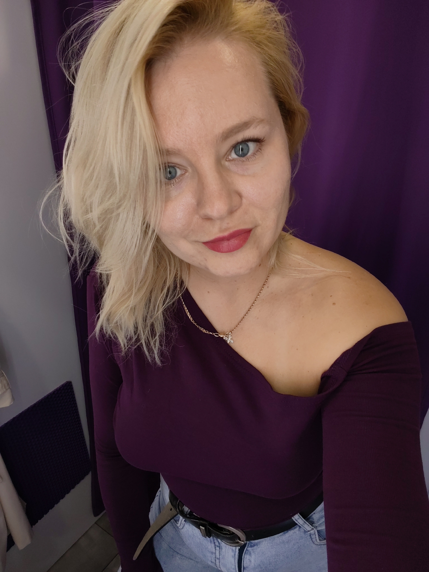 A blonde woman with blue eyes and pink lipstick is looking at the camera in a selfie. She is wearing an off-the-shoulder deep purple top, blue jeans, and a black belt with a silver buckle. A gold necklace with a small pendant rests on her décolletage. The background is a dark purple curtain. The lighting suggests an indoor setting, possibly a dressing room. The image does not contain information to determine its specific location within Izhevsk, Russia.