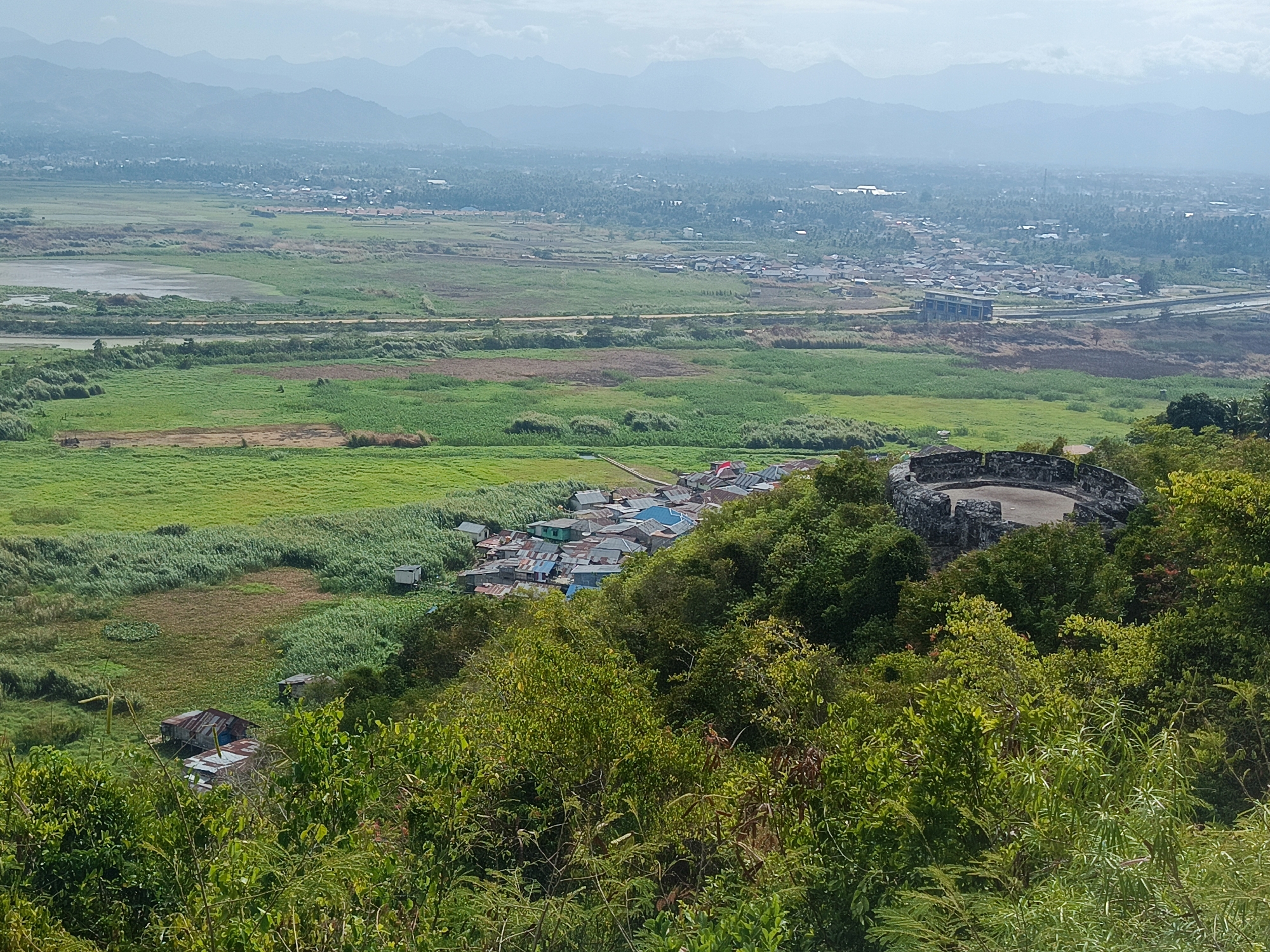 The image is a landscape view of a settlement in Gorontalo, Indonesia. In the foreground, lush green foliage and trees dominate, with a weathered stone structure, possibly a historical monument or observation deck, visible on the right. The middle ground features a cluster of houses with various colored roofs, nestled amongst the trees. Beyond this, a vast expanse of green fields and marshland stretches towards a distant town or city. The town is characterized by numerous buildings and structures, with a prominent multi-story building visible. In the background, hazy mountains form a backdrop under a pale sky. The scene appears to be captured during daylight, with soft lighting conditions.
