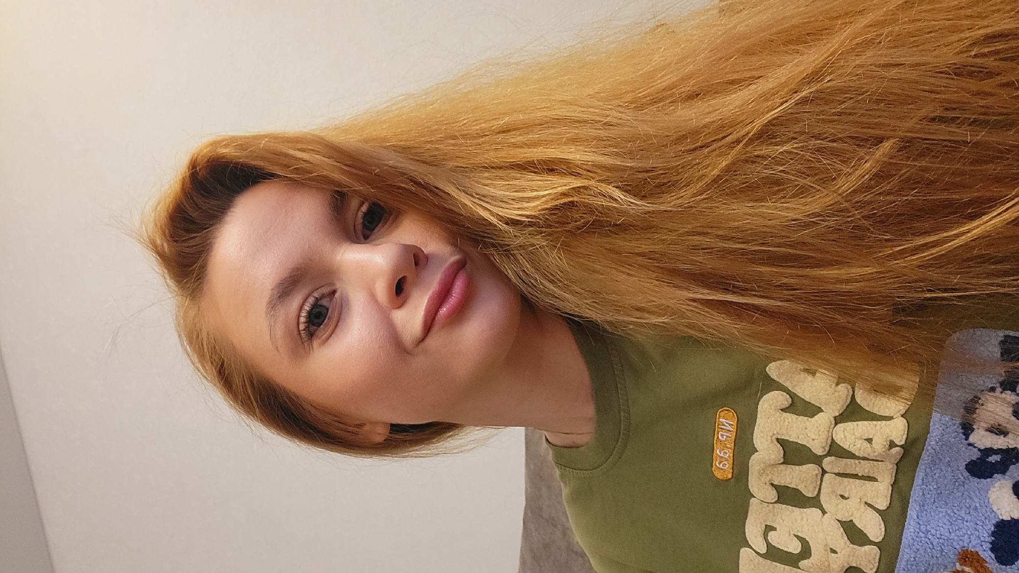 A close-up selfie photograph shows a young woman with fair skin and long, light brown to strawberry blonde hair. The hair is voluminous and partially covers the upper portion of the image. Her eyes are green and she has a slight pout. She is wearing a green t-shirt with a graphic design. The background is a plain, off-white wall on the left side. A patterned material is visible on the lower right edge. The exact location in Chaikovsky, Russia, cannot be determined from the image.