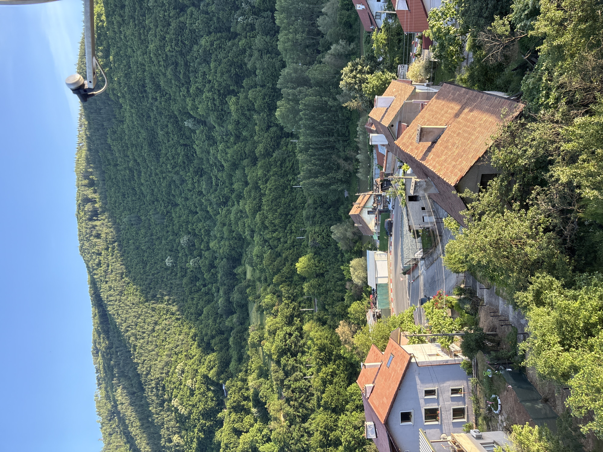This is an aerial view of a village nestled at the base of a lush green mountain, likely in Redina, Bulgaria, given the context. The scene is dominated by a steep, densely forested mountainside that slopes down towards a small settlement. The sunlight casts long shadows across the mountain, highlighting the texture of the dense trees.
The village itself consists of several houses with terracotta tiled roofs, interspersed with areas of dense green foliage. In the foreground, a light blue house with multiple windows and a grey, corrugated metal balcony is visible on the left. Adjacent to it, a pathway with dark metal railings leads down into a garden area. Further into the village, other houses with similar red tile roofs are scattered.
A road runs through the village, with a few parked cars and some small industrial or storage buildings visible. A traffic barrier and some fencing are present near the road. The overall impression is one of a quiet, rural community situated in a picturesque natural setting. The sky is a clear, bright blue. In the upper right corner of the frame, a metal pole with a satellite dish is visible, extending from an unseen structure.