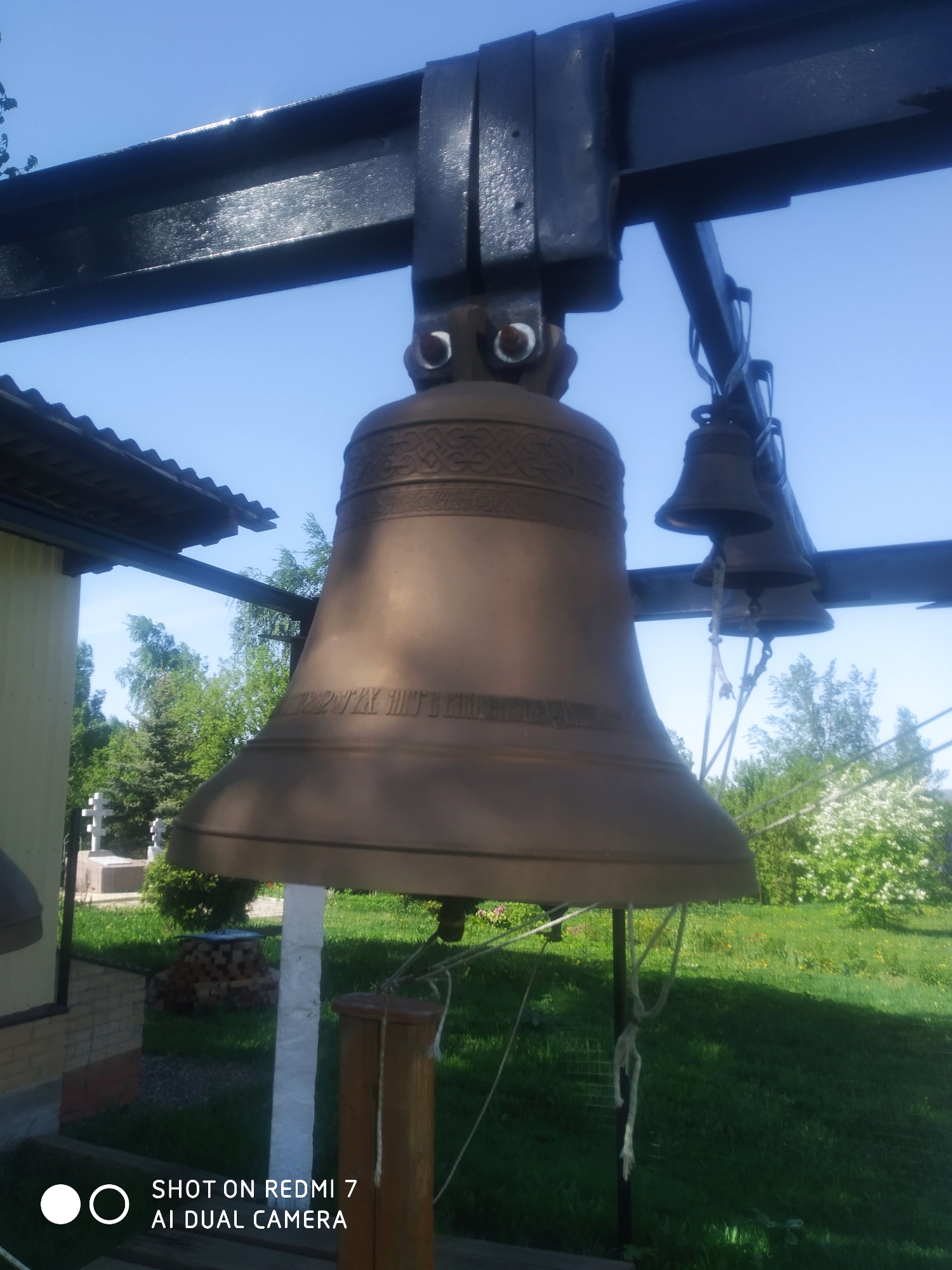The image depicts a large bronze bell suspended from a black metal frame, likely a bell tower or structure for ringing. Smaller bells hang to the right of the larger bell. The bell is outdoors in what appears to be a grassy area with trees and a building visible in the background. The photo was taken in Ochansk, Russia.