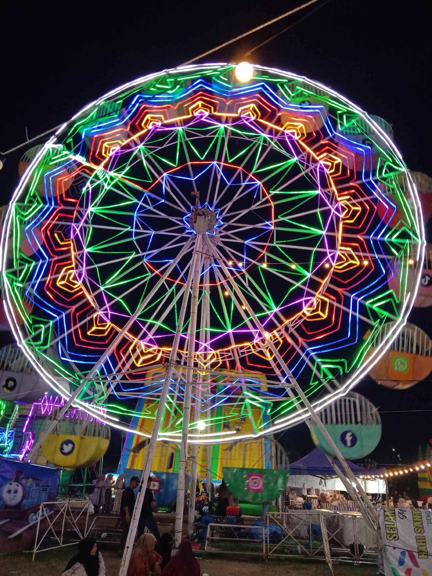 The image captures a night scene in Geulumpangsulu Timu, Indonesia, dominated by a brightly illuminated Ferris wheel. The wheel is decorated with concentric rings of various neon colors, including green, blue, purple, orange, and red, creating a vibrant display against the dark sky. The structure is supported by white metal beams. Gondolas are partially visible, some featuring social media logos such as Twitter, Facebook, Instagram, and WhatsApp.
At the base of the Ferris wheel, several people are seated or standing. Some are wearing head coverings. Behind the Ferris wheel is a partially visible building with striped walls. To the right, a string of lights illuminates a concession stand with vendors and additional onlookers. A sign is visible near the front right. The ground appears to be grass or dirt.