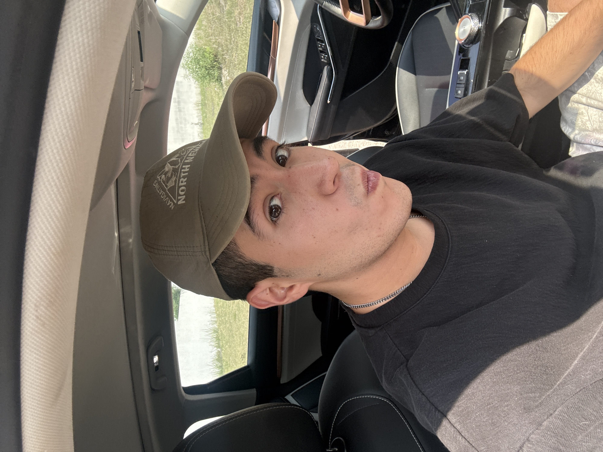 A young man is inside a car. He's wearing a dark t-shirt and a baseball cap with text and an image on it. The interior of the car features black leather seats with white stitching, a black dashboard, and a silver gear shift console. Outside the car window, there is green foliage visible. The location is not able to be determined as San Bernardino Tlaxcalancingo, Mexico from this image.
