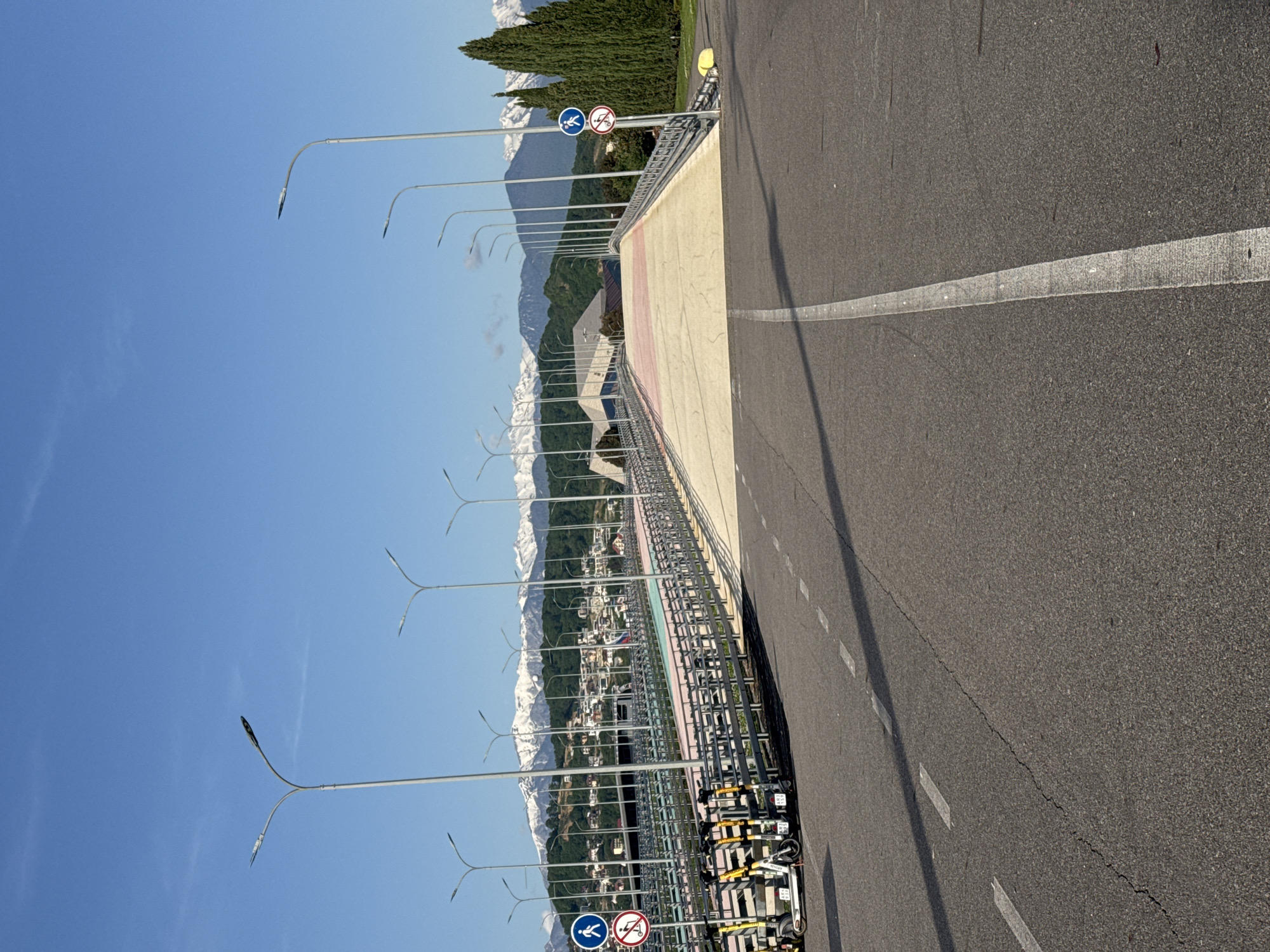 The image depicts a bridge in Sirius, Russia, with a view of snow-capped mountains in the background. The bridge has a pedestrian walkway with metal railings and is lined with modern streetlights. A pedestrian crossing sign and a no-scooter sign are visible at one end. The road on the bridge has a white dividing line. Several scooters are parked on the walkway near the signs. The sky is clear and blue. The image suggests a bright, sunny day.