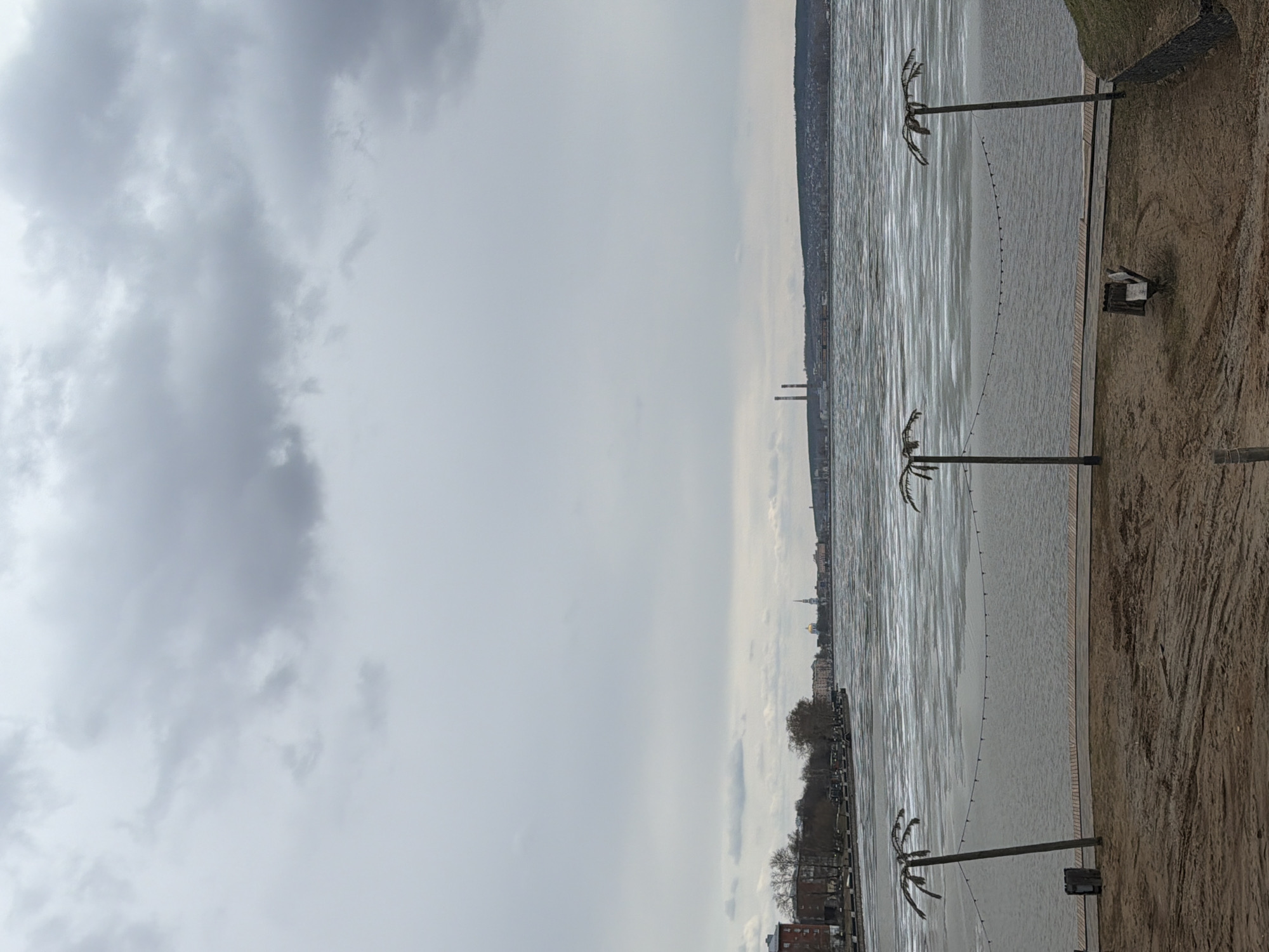 This image presents a wide, horizontal landscape view of a waterfront scene in Votkinsk, Russia, under a heavily overcast sky. The lighting suggests it is daytime, likely during a cold period given the visible ice on the water.
In the foreground, a sandy or muddy bank runs along the right side, bordered by what appears to be a wooden walkway. Three decorative, artificial palm tree-like structures, fashioned from dark metal with sparse, spiky fronds, are spaced along this bank. A string of small lights forms an arc between two of these "palm trees," extending out over the water. A small, dark wooden bench or similar structure is situated on the bank near the center.
The midground is dominated by a large body of water, presumably the Votkinsk Reservoir. Its surface is partially covered with sheets of ice or frozen patterns, reflecting the grey sky and giving it a textured, muted appearance.
Across the water, the distant shoreline reveals a mix of natural and urban landscapes. On the far left, hills covered with what appear to be bare trees rise from the water's edge. As the view progresses to the right, a city skyline comes into focus, featuring various buildings. Two prominent industrial smokestacks or towers are visible in the middle distance, and further to the right, a distinctive golden-domed church spire or cupola can be seen against the horizon.
The sky is a uniform, heavy grey, filled with thick, diffused clouds, contributing to a tranquil and somewhat somber atmosphere. No people or specific activities are depicted, portraying a quiet, expansive panorama. The artificial palm trees and lights add an unexpected, decorative element to the otherwise natural and developed landscape. No text is visible within the image.