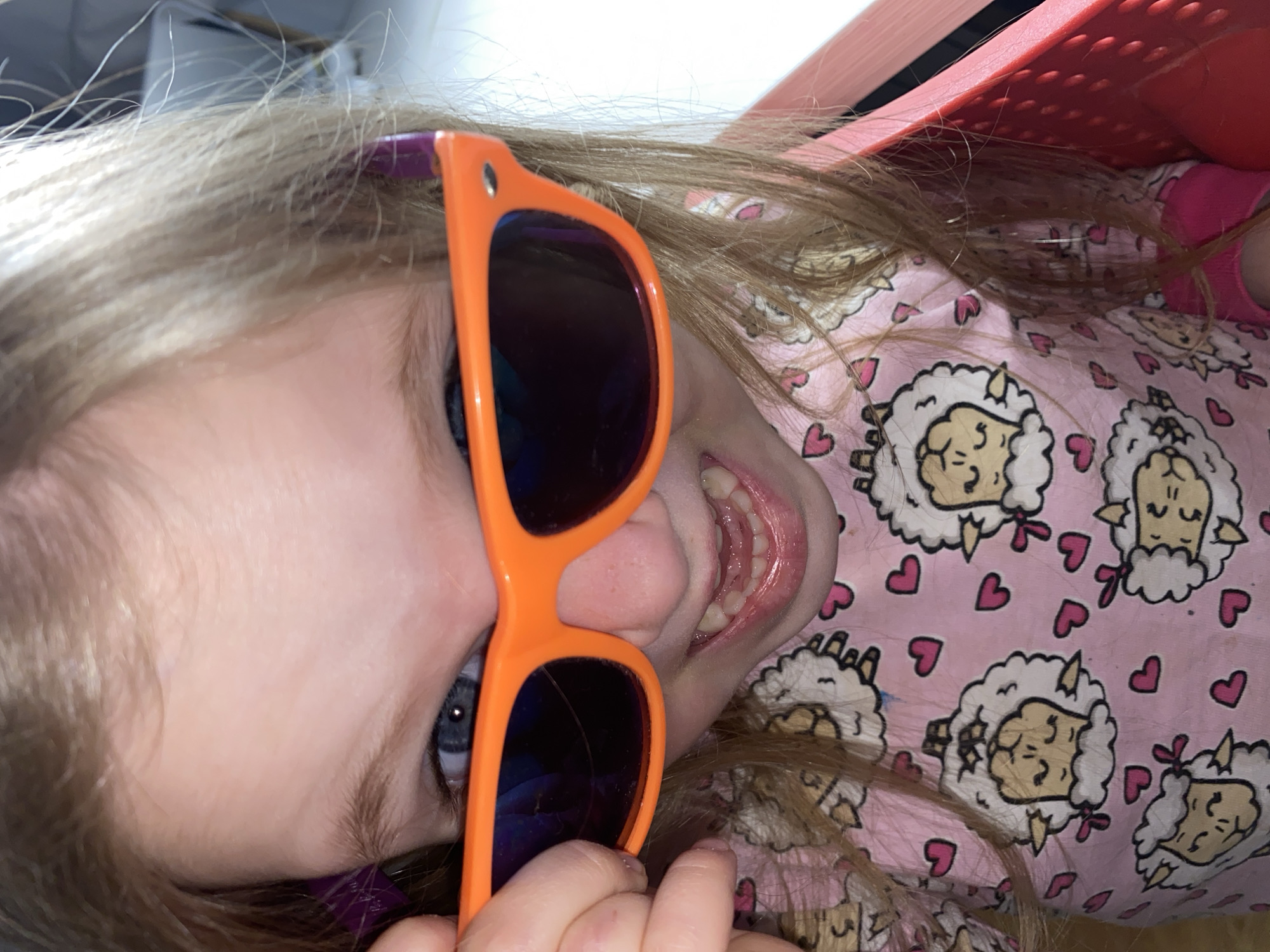A young child lies on their side, facing the camera, wearing sunglasses with orange frames and dark lenses. The child is wearing a pink pajama top with a pattern of sheep and hearts. They are positioned near a red object. The location is unknown.