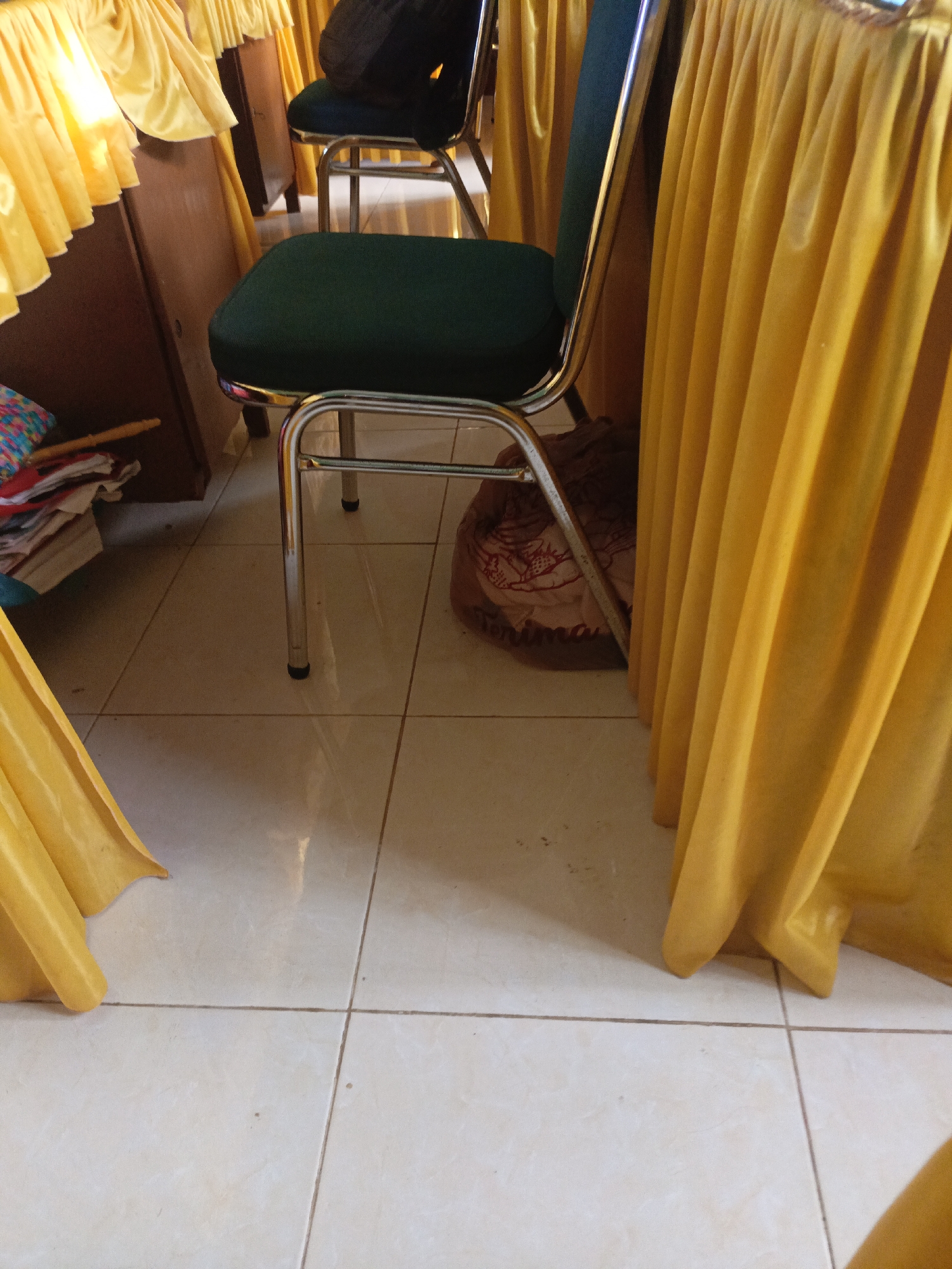 A close-up, medium-angle shot shows the legs and seats of two dark green upholstered chairs with shiny metal frames, positioned on a tiled floor. The chairs are partially obscured by bright yellow tablecloths that drape down the sides. To the left, a stack of colorful folded items and a wooden object are visible behind a dark wooden cabinet. The lighting suggests an indoor setting, possibly a room in Lhokseumawe, Indonesia, used for events or gatherings. The floor tiles are a light cream color with visible grout lines.