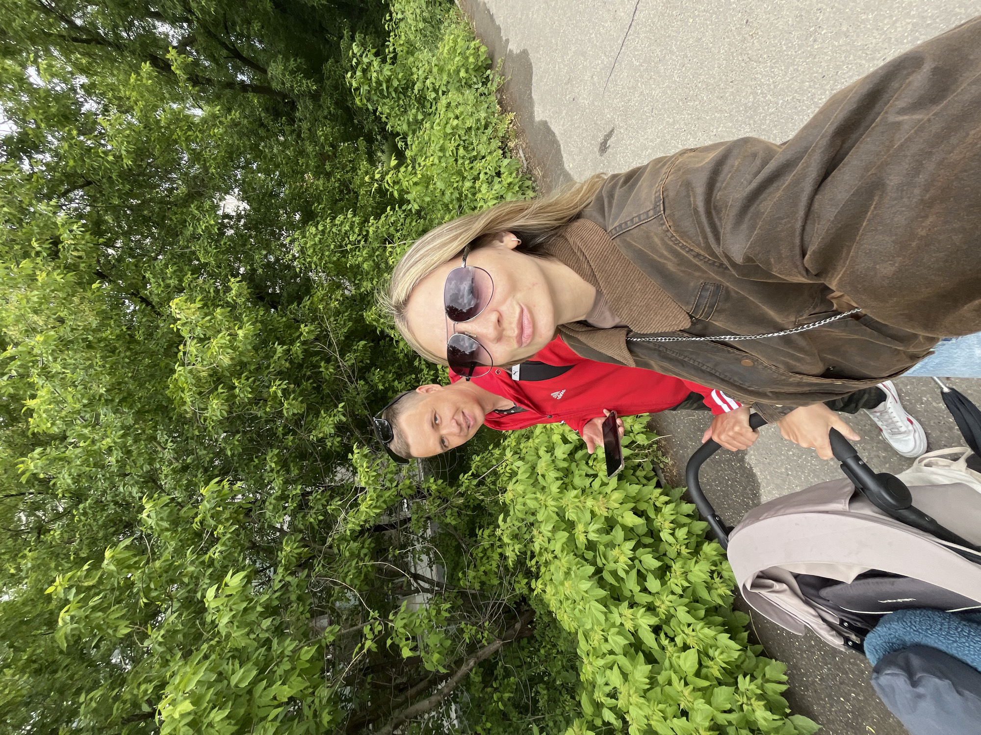 A medium-shot selfie taken on an overcast day shows a woman wearing a brown waxed-canvas jacket and sunglasses, pushing a baby stroller. A man is behind and to the left of her, wearing a red jacket with white stripes on the sleeves and holding a cell phone. They are standing on a paved path adjacent to dense green foliage. The location is possibly in Chaikovsky, Russia.