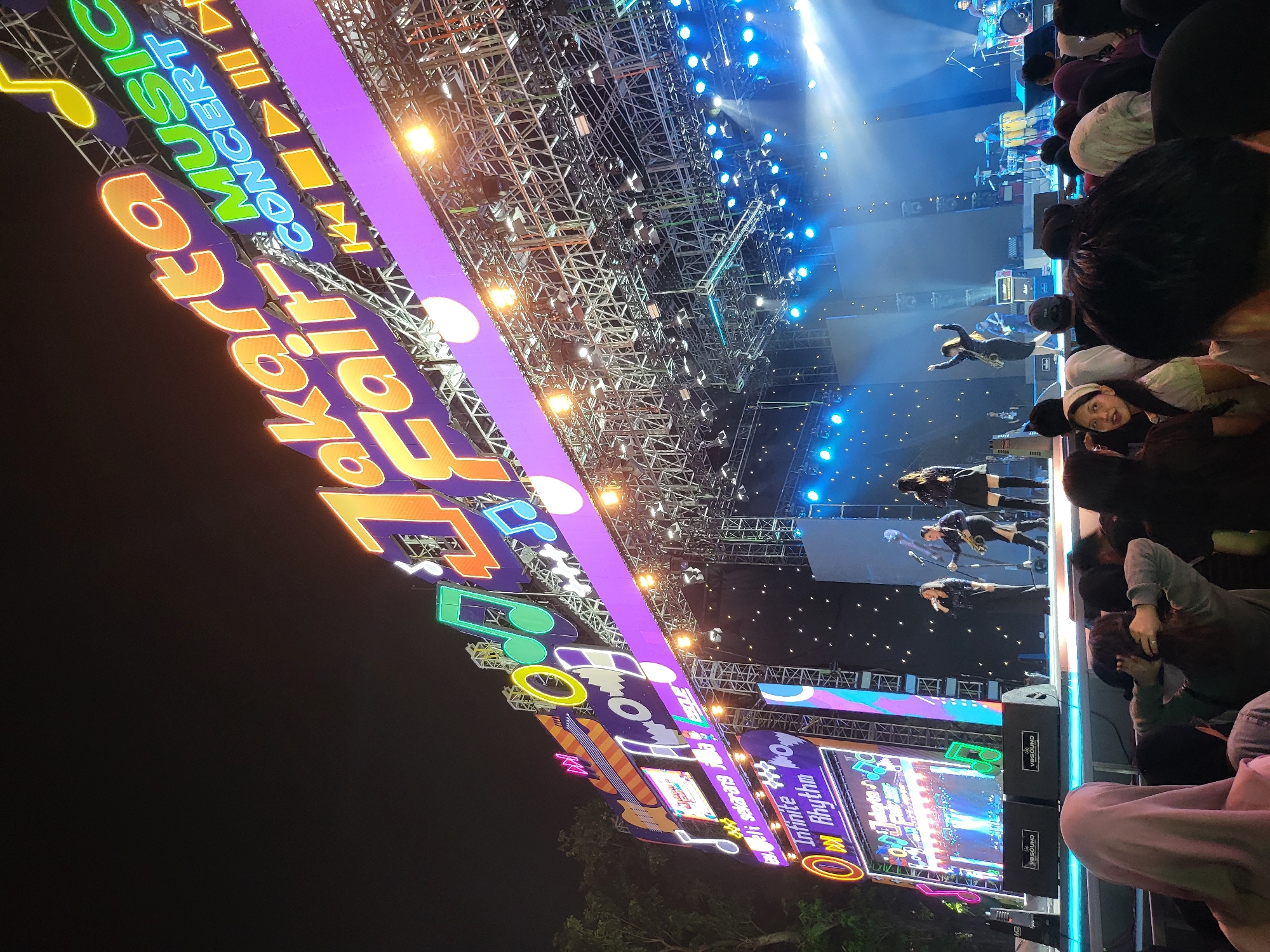 This image captures an outdoor nighttime concert at the Jakarta Fair in North Jakarta, Indonesia. A large, brightly lit banner overhead reads "Jakarta Fair Music Concert" in large, colorful neon letters. Below the banner, a stage is set up with performers, a drum kit, and other musical instruments. Bright stage lights illuminate the performers and the surrounding area, casting a vibrant blue and white glow. A crowd of people is gathered in front of the stage, watching the performance. The overall atmosphere appears to be energetic and festive.