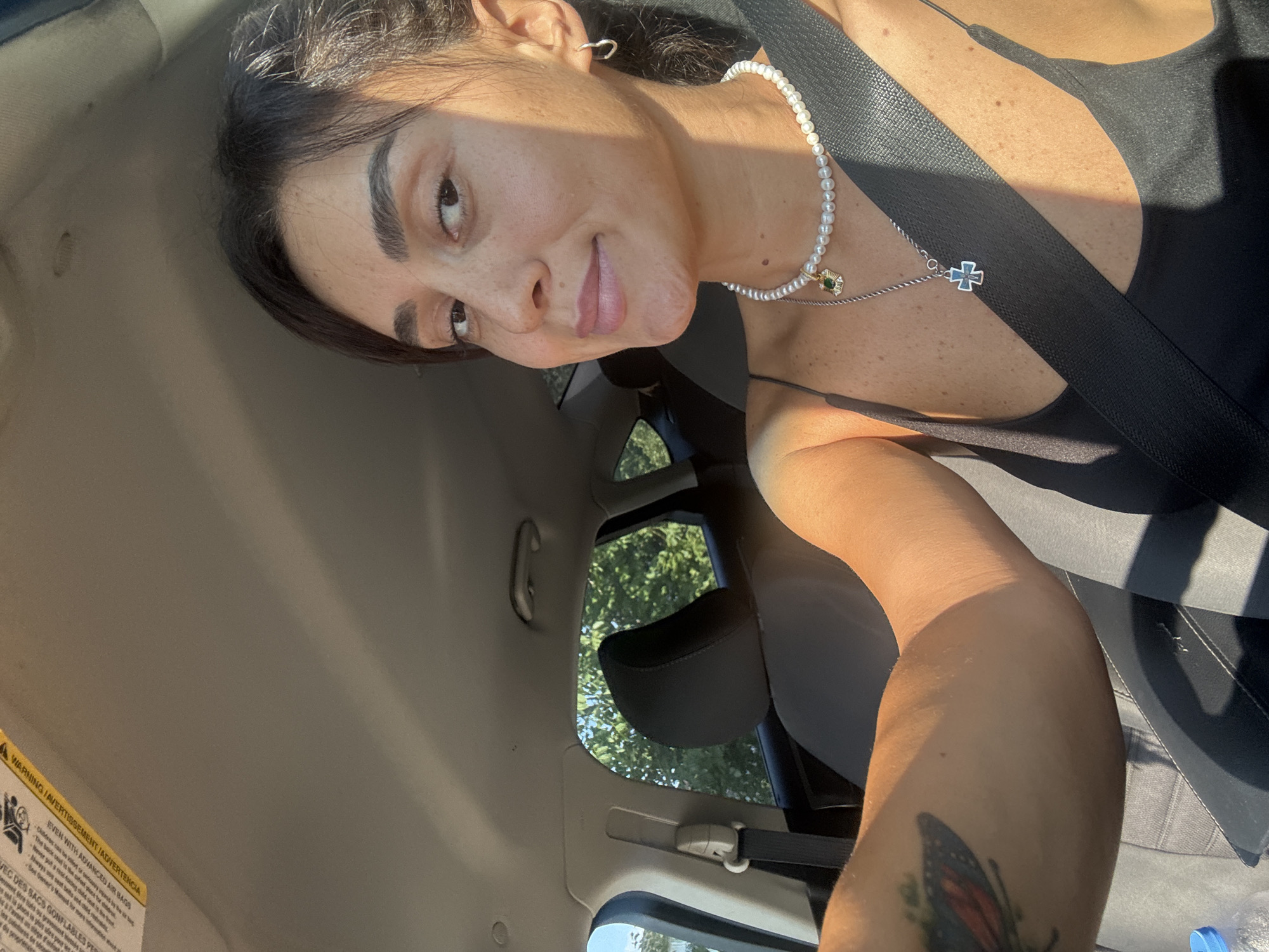 A young woman with dark hair is taking a selfie in a car. She is wearing a black tank top, a pearl necklace with a green pendant, and a silver cross necklace. She has a butterfly tattoo on her left arm. The car appears to be driving through a tree-lined area, possibly a suburban neighborhood or a park. The sun is shining brightly, casting shadows inside the car. The woman is looking at the camera and smiling. There is no discernible information in the image to suggest a location in Tbilisi, Georgia.