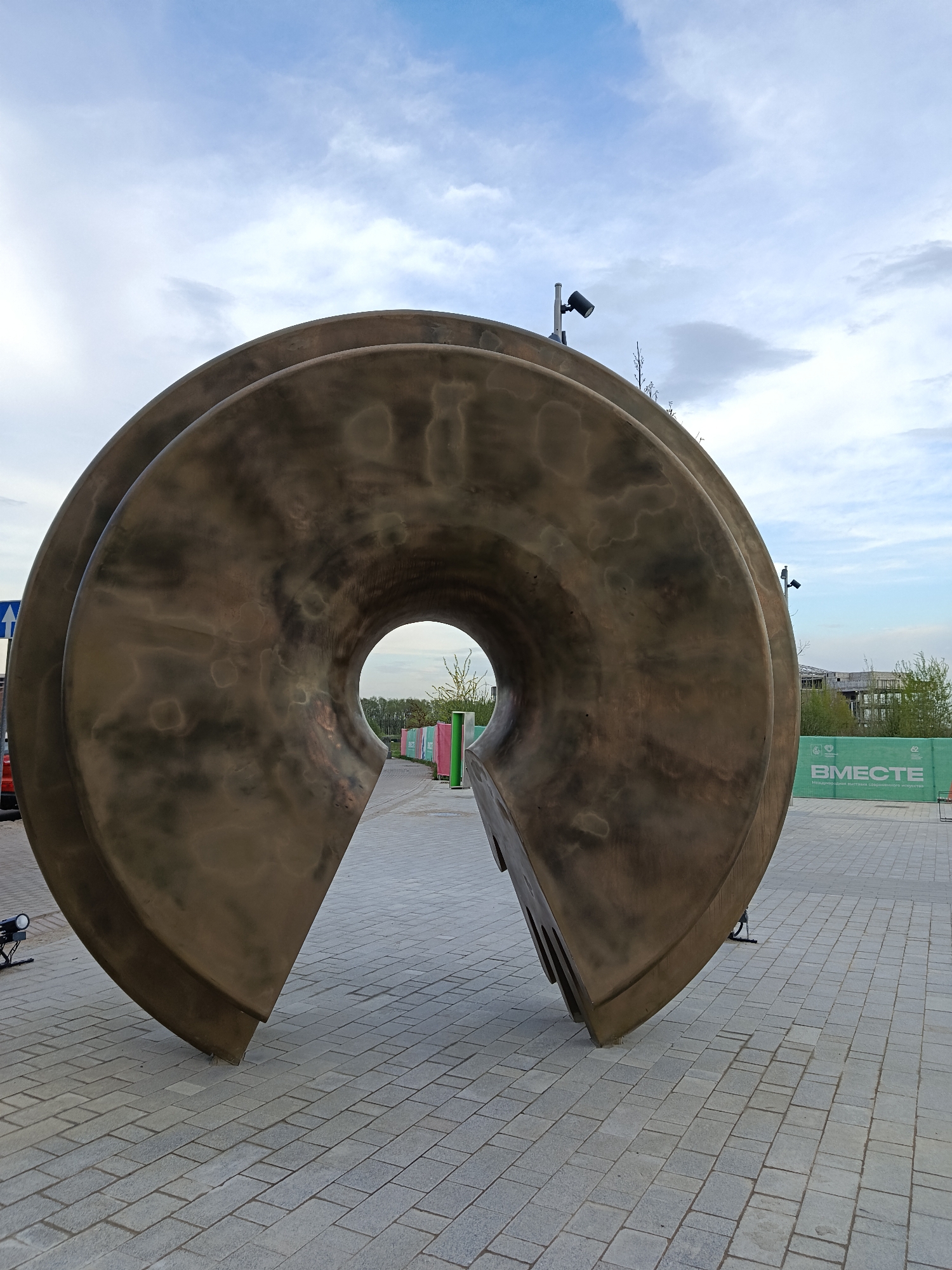 A low-angle, full shot depicts a large, abstract sculpture in a public area, likely a plaza or park. The sculpture is made of a dark, weathered bronze material and consists of two large, concentric, semi-circular forms that are slightly offset from one another. The sculpture is split open, creating a circular opening in the center, through which glimpses of trees and buildings can be seen.
The ground surrounding the sculpture is paved with gray bricks. In the background, there is a green fence with white lettering that says "ВМЕСТЕ" in Russian. The sky is partially cloudy with a mix of blue and white.
The location of the photograph is in Moscow, Russia.