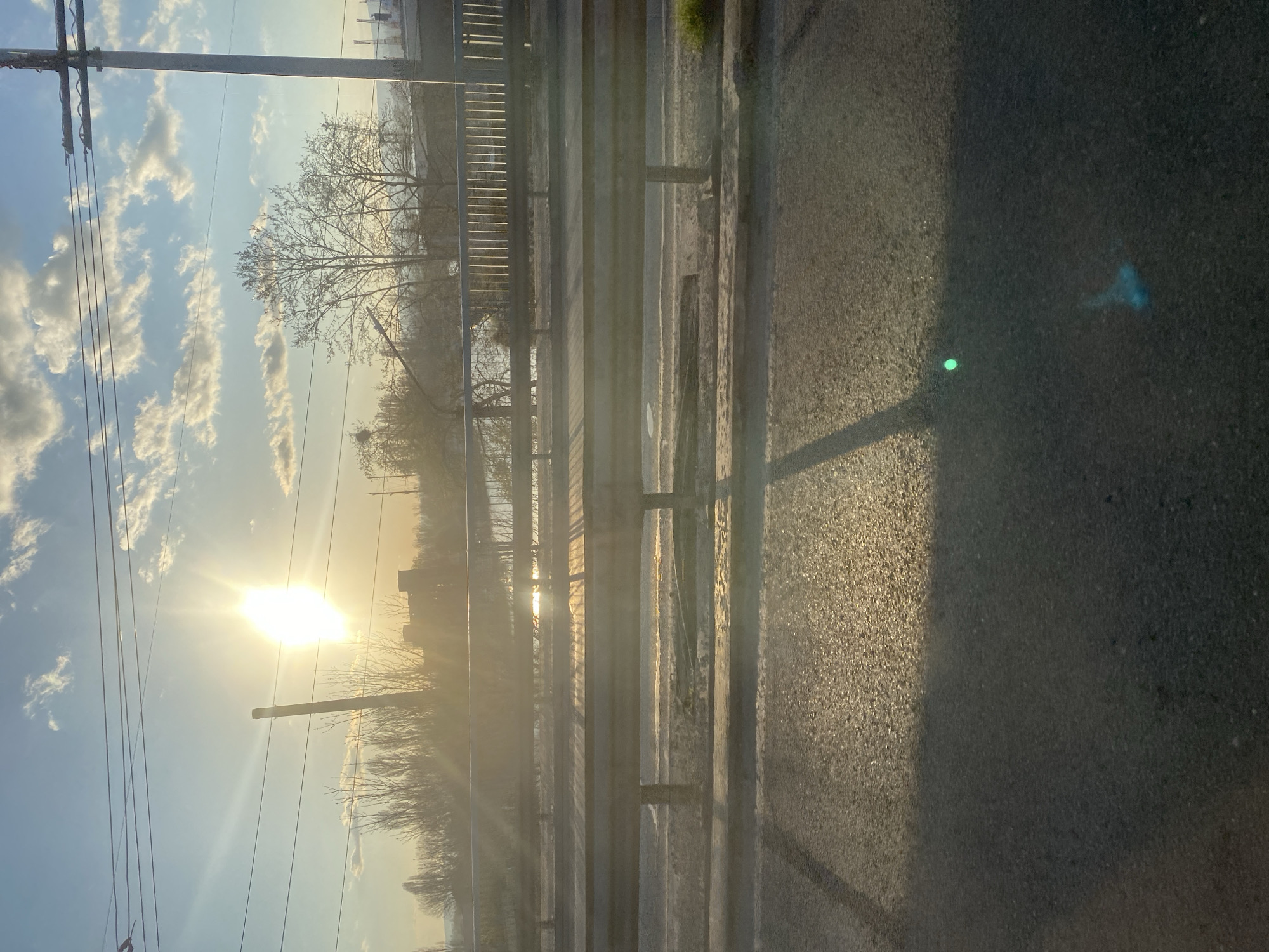 The image shows a view from a bridge or elevated structure, potentially in Varna, Bulgaria, although there's nothing definitively identifying it as such. A metal railing runs horizontally across the frame. Beyond the railing, the sun is low on the horizon, partially obscured by buildings and trees. Silhouetted trees and structures are visible. Power lines run vertically along the left side of the image. The asphalt surface of the bridge is visible on the right, with sunlight and shadows cast across it. There are no people visible in the image.