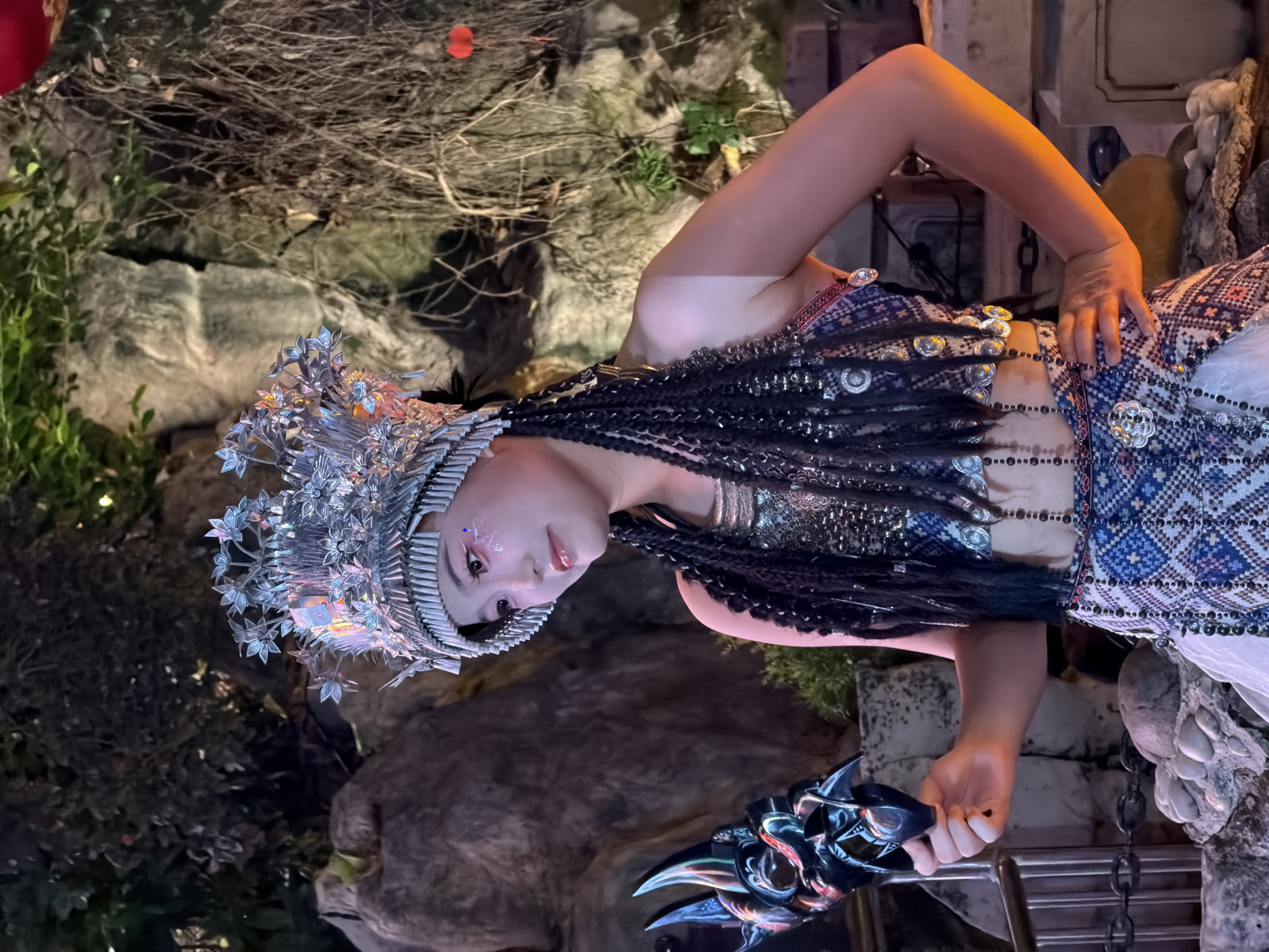 A person in elaborate costume, adorned with a large, intricate silver headdress and braided hair, is posed in an outdoor setting at night. The costume is detailed with colorful woven patterns and metallic embellishments. The individual holds a dark, sharp object with a decorative, possibly monstrous, design in their right hand. The background features natural elements like rocks and foliage, with artificial lighting creating a dramatic effect. The overall atmosphere suggests a performance or staged event, possibly related to a festival or cultural celebration. The person's makeup is striking, with glitter accents on their eyes. The scene is illuminated by artificial lights, with no indication of the weather or specific time of day beyond it being nighttime. The location is identified as Yuzhong District, China.