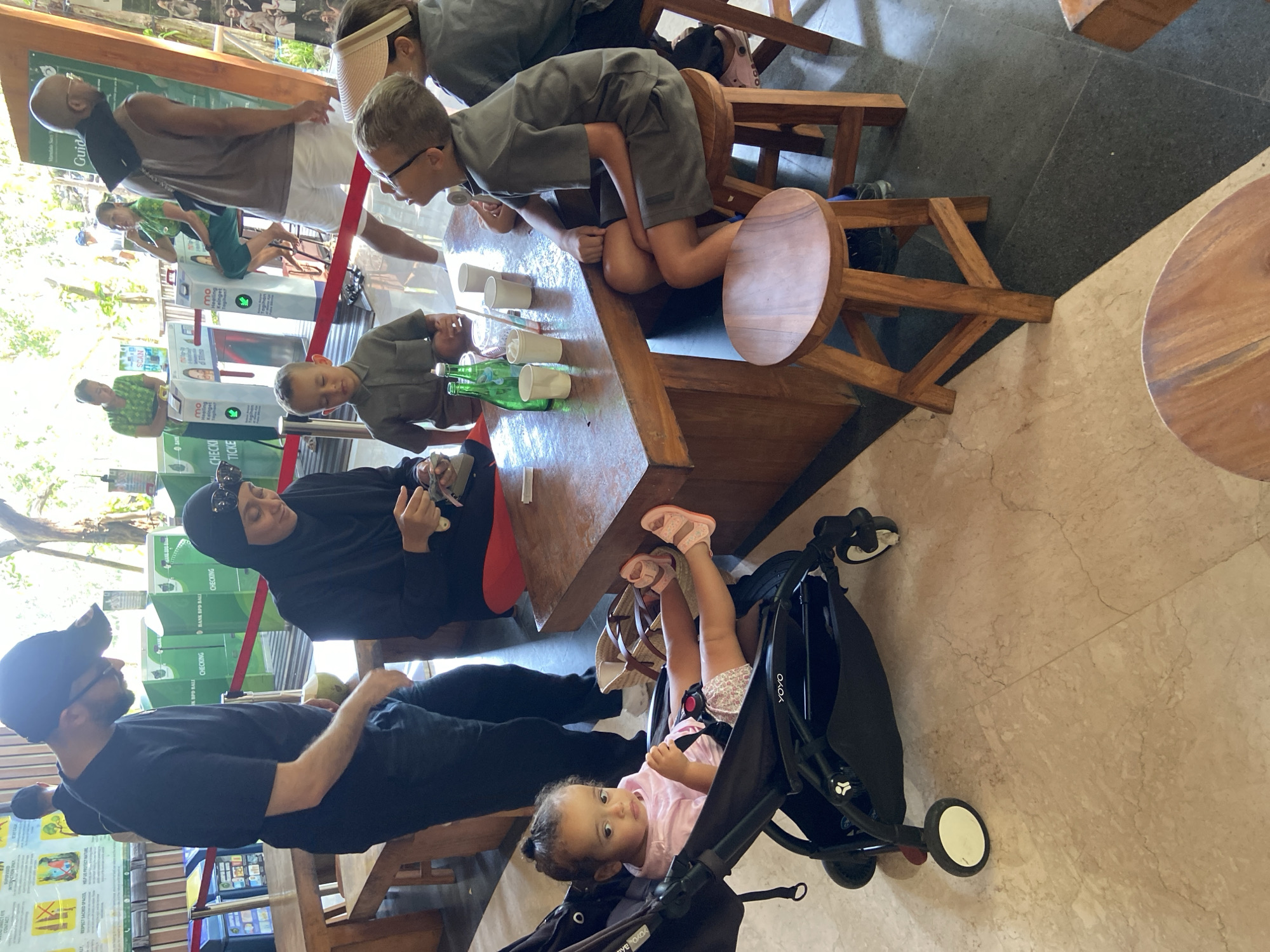 In Ubud District, Indonesia, at a café or restaurant adjacent to an entrance with ticket barriers, a group of people are seated at a wooden table. A man in a black t-shirt and a black cap stands to the left, looking down. A woman wearing a black hijab and sunglasses is seated at the table, holding small items in her hands. A young boy in a grey shirt is also seated at the table, looking down. A baby is in a black stroller to the left of the table. Two children are seated on wooden stools at the right side of the table. The environment appears to be an indoor seating area with wooden furniture and a marble floor. The background shows green ticket barriers and foliage, suggesting an outdoor element beyond the seating area.