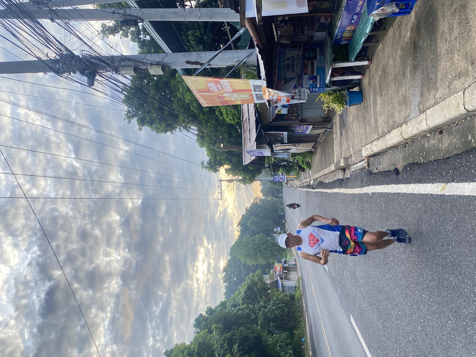 A man is running on a paved road, seen from the chest down, wearing a white tank top with blue trim and a logo that reads "DUC 50 TAGUM DAVAO", and brightly colored shorts. He is wearing a white cap and dark athletic shoes. He is running on the right side of the road, in the direction of the viewer. The road is bordered by a concrete sidewalk on the right, with a row of small shops and food stalls. A large, yellow banner with red text is hanging from one of the stalls. Behind the stalls, there are utility poles with a dense network of wires. Further down the road, there is a motorcycle with a rider. The left side of the road is lined with lush green trees and foliage. The sky is cloudy with patches of sunlight breaking through, suggesting either dawn or dusk. The scene is likely in Davao City, Philippines, as indicated by the text on the runner's shirt.