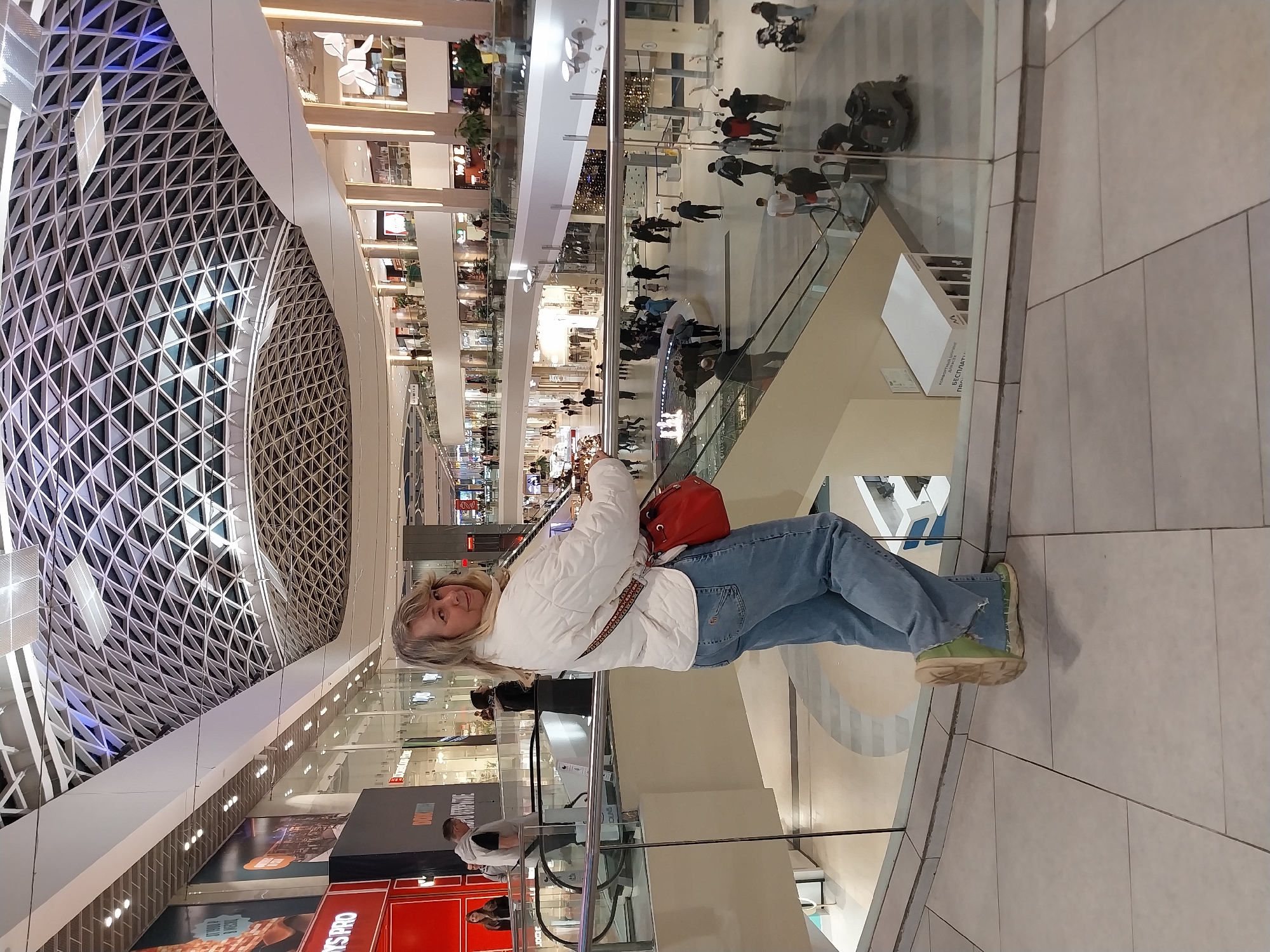A woman in a white jacket, blue jeans, and green sneakers with a red backpack stands inside a multi-story shopping mall. The mall is in Sochi, Russia. The mall has a distinctive domed roof with a geometric pattern and is filled with stores and people on multiple levels. The woman is standing on an upper level with a glass railing, overlooking the activity below.